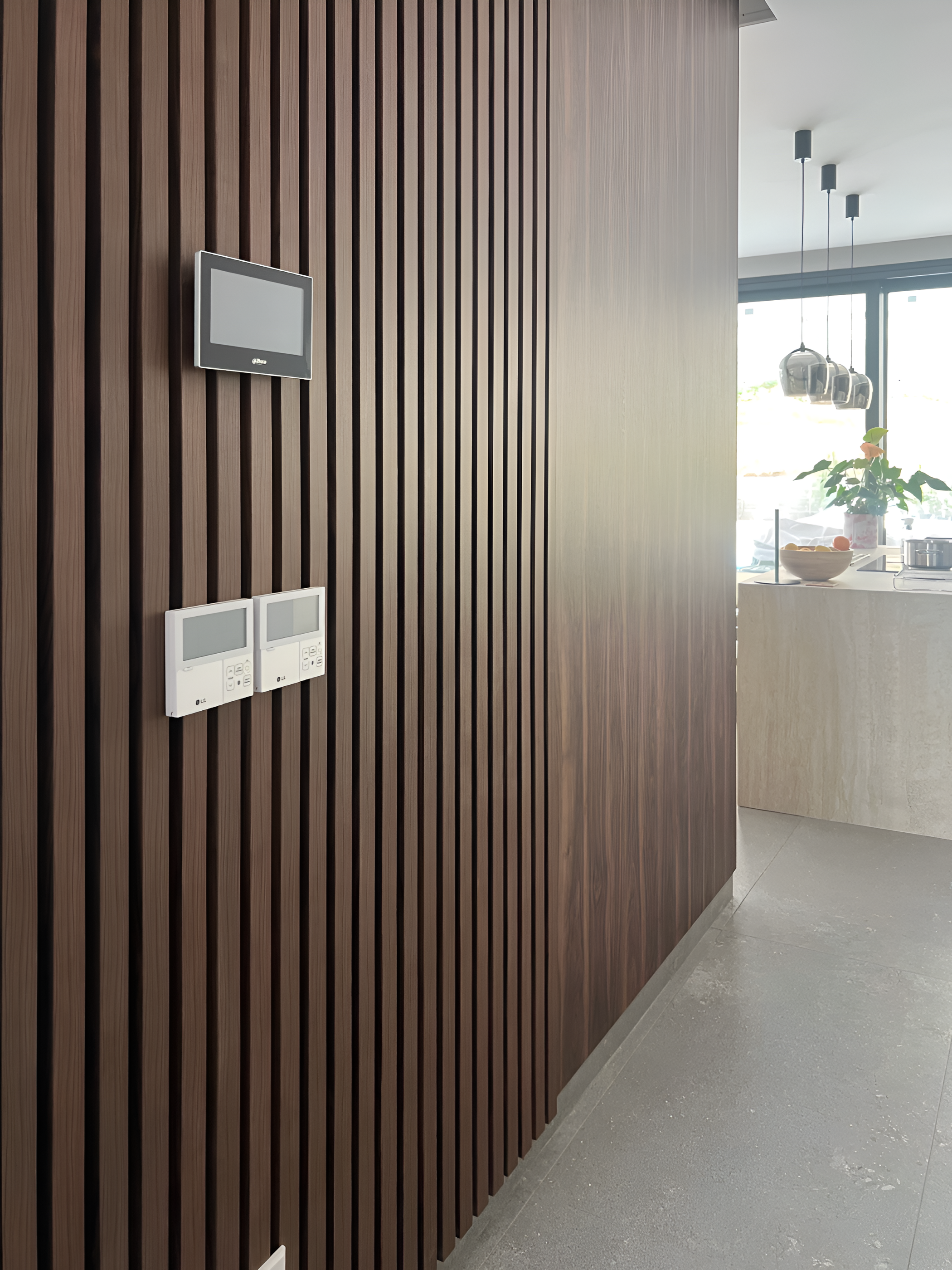 Wood-paneled wall with two black wall-mounted screens and three white thermostats. Part of a modern kitchen with a white countertop, potted plant, and hanging pendant lights in the background.