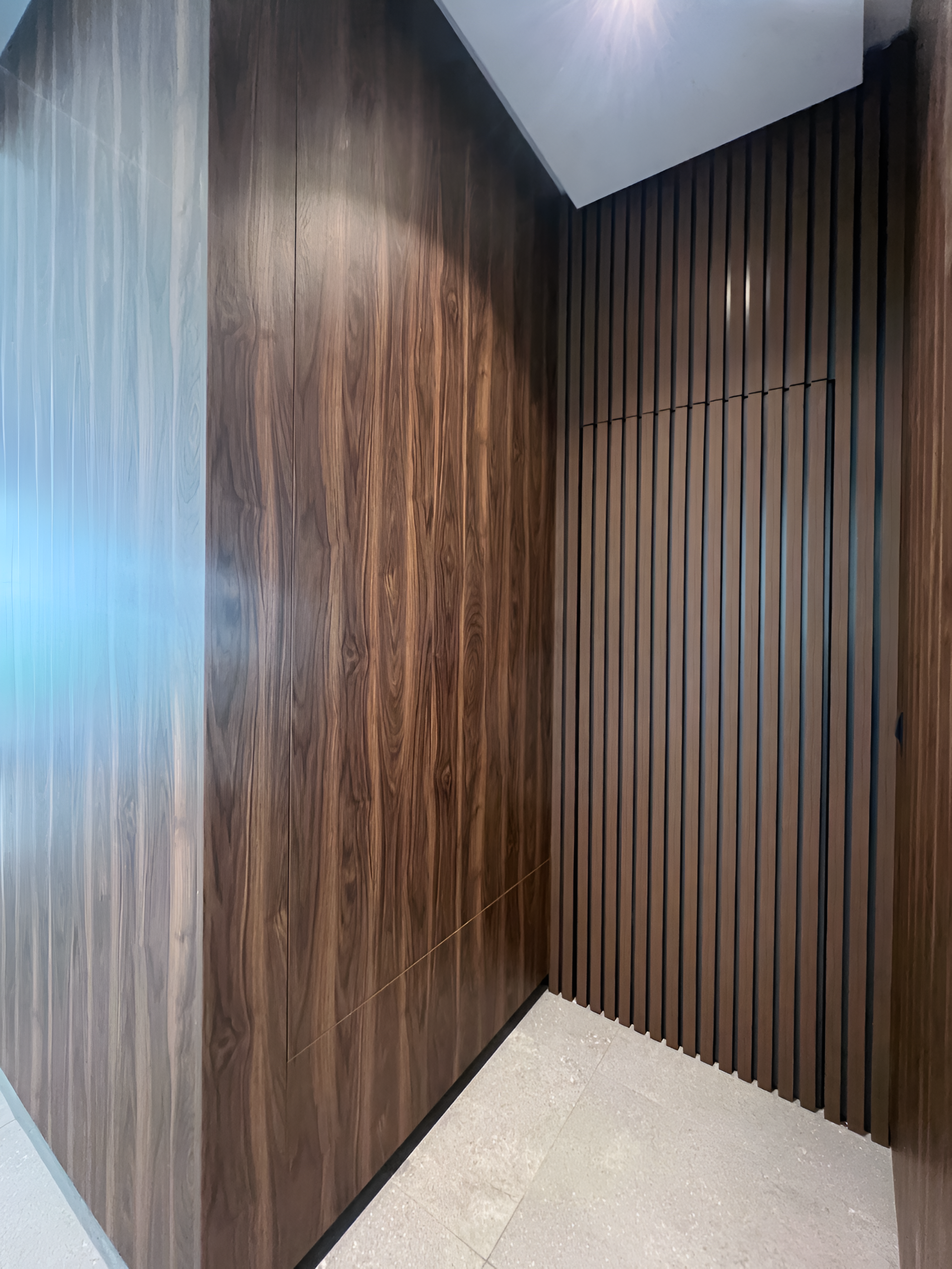 View of an interior corner with wood-paneled walls, one with horizontal slats and the other with a smooth finish, and a light-colored tiled floor.