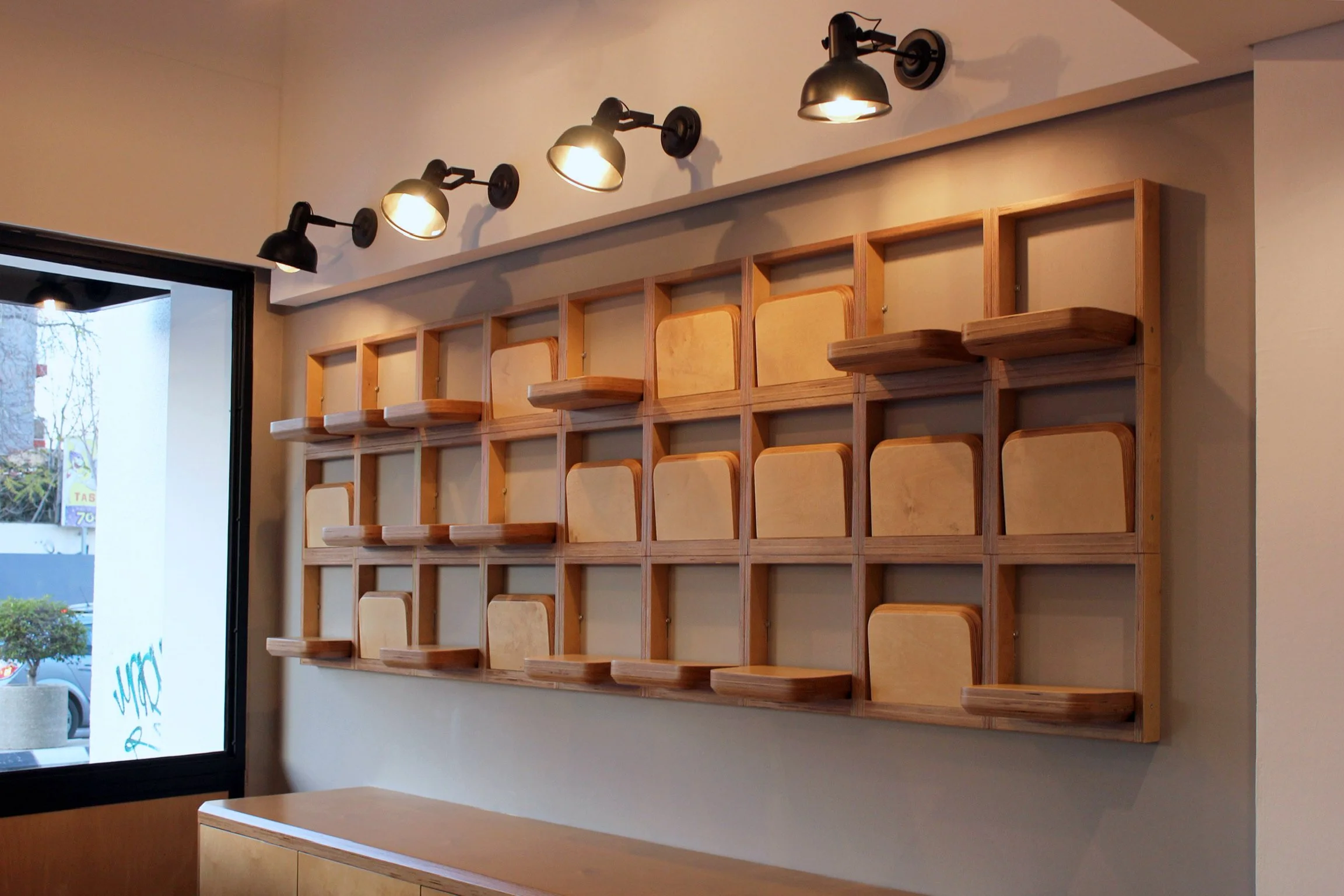 Interior of a store or cafe with a wooden cubed shelf on the wall, black industrial-style light fixtures above, and a partially visible window showing outdoor trees and signage.