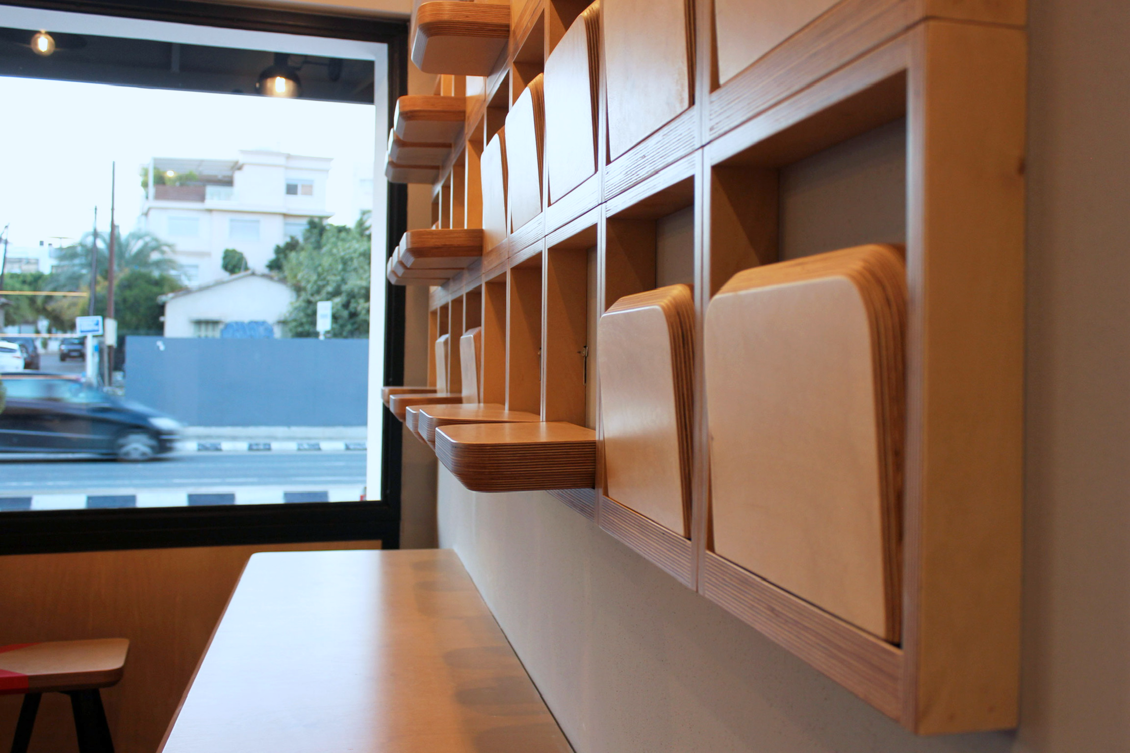 Interior of a cafe or restaurant with wooden shelves on the wall and a window showing a street scene outside.
