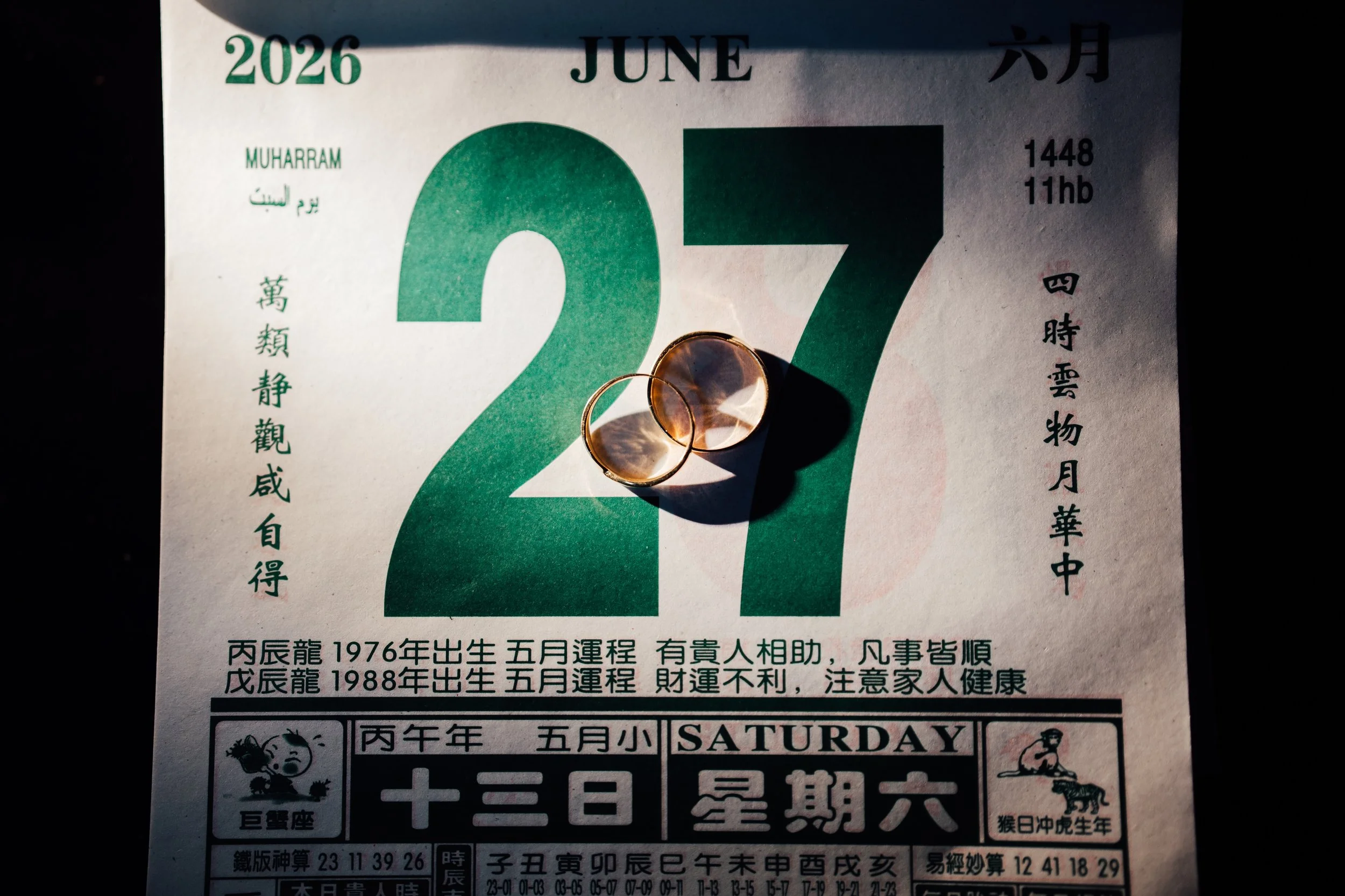 A calendar page for June 2026 displayed with a pair of wedding rings placed on it. The calendar includes Chinese characters and some English text, with the date 27 prominently shown. The rings cast a shadow on the paper.
