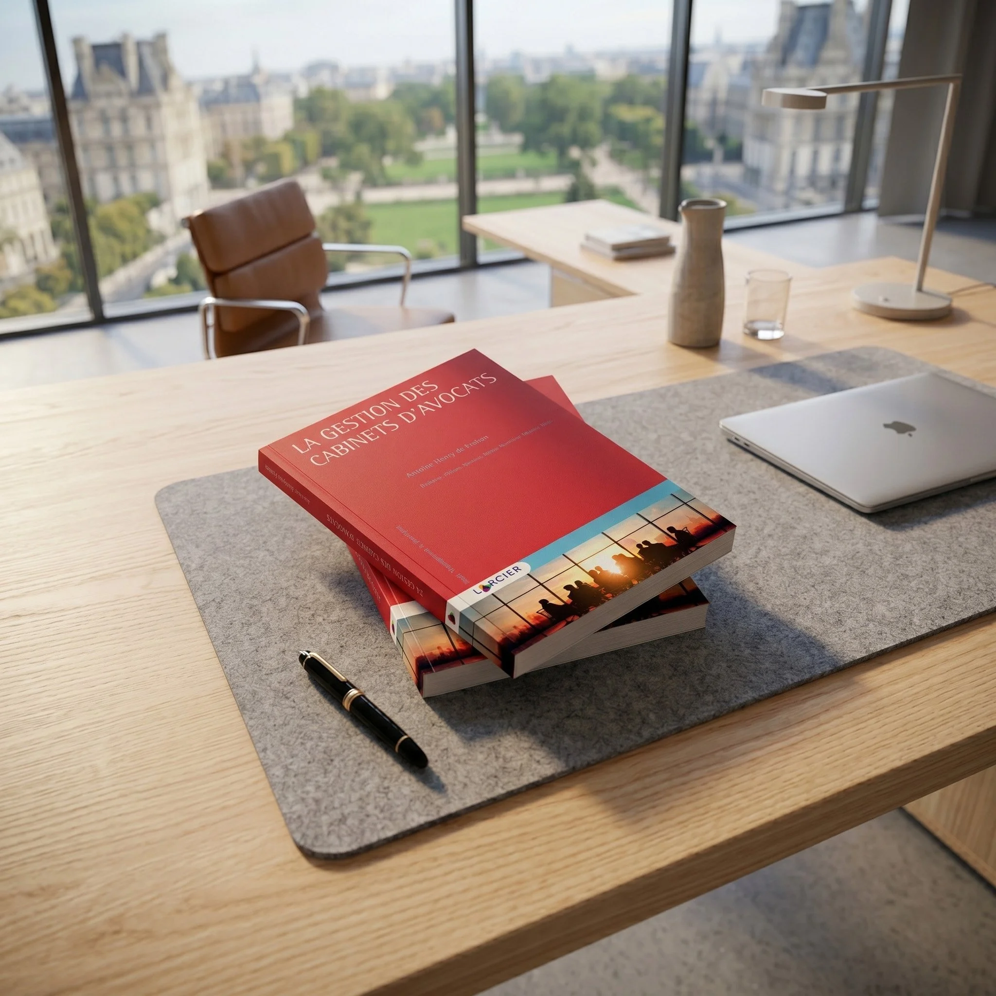 An office space with a large window view of a city park and historic buildings. A wooden desk holds a closed laptop, a pen, a red book, a white ceramic pitcher, a glass of water, and a modern desk lamp. A brown leather chair with metal arms is positioned at the desk.