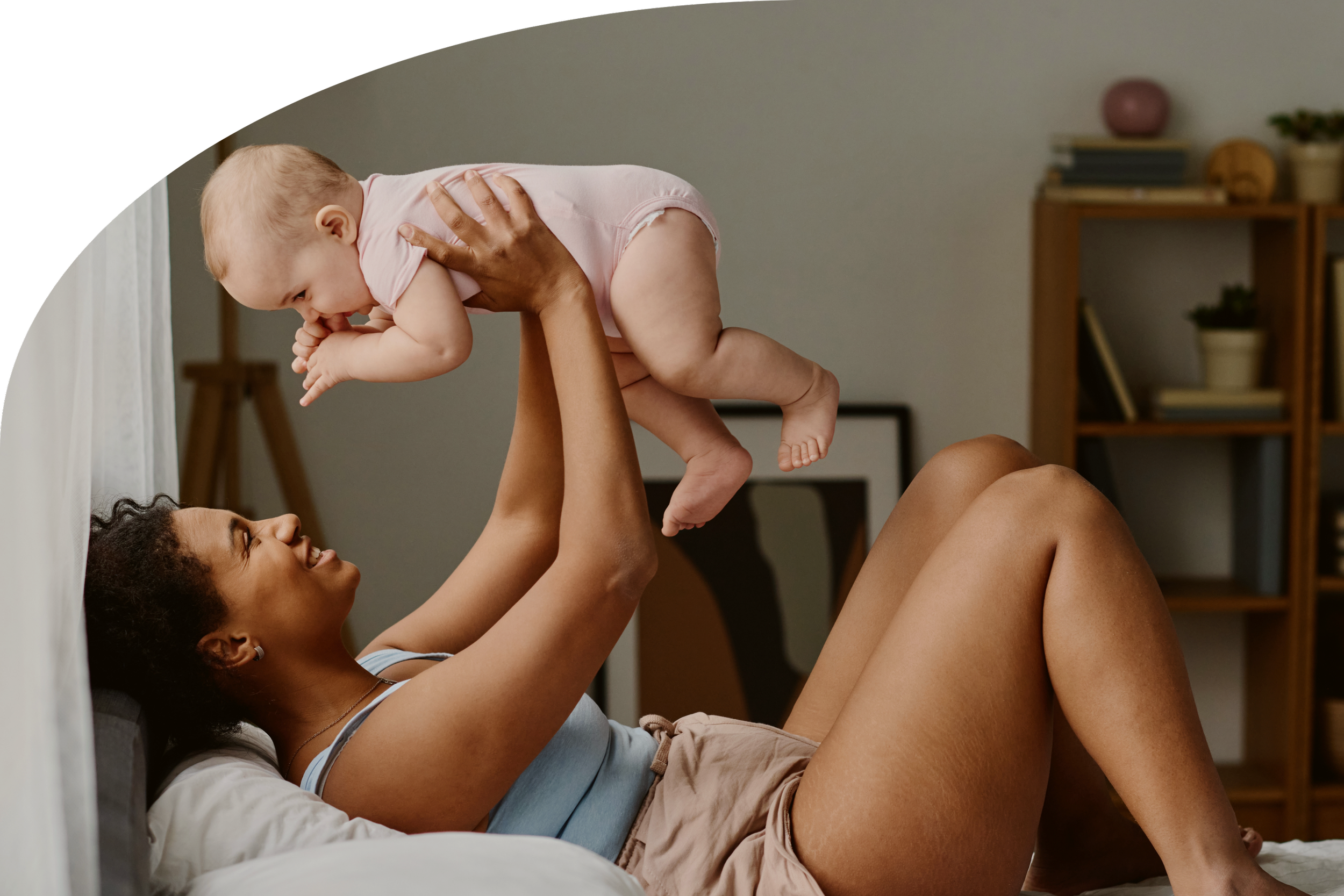 A woman lying on a bed holding a baby and smiling. The photo lies within a frame that has 1 curved edge. This photos represents Strong Roots OT where we provide maternal health & pediatric occupational therapy services for women & kids in south NJ.