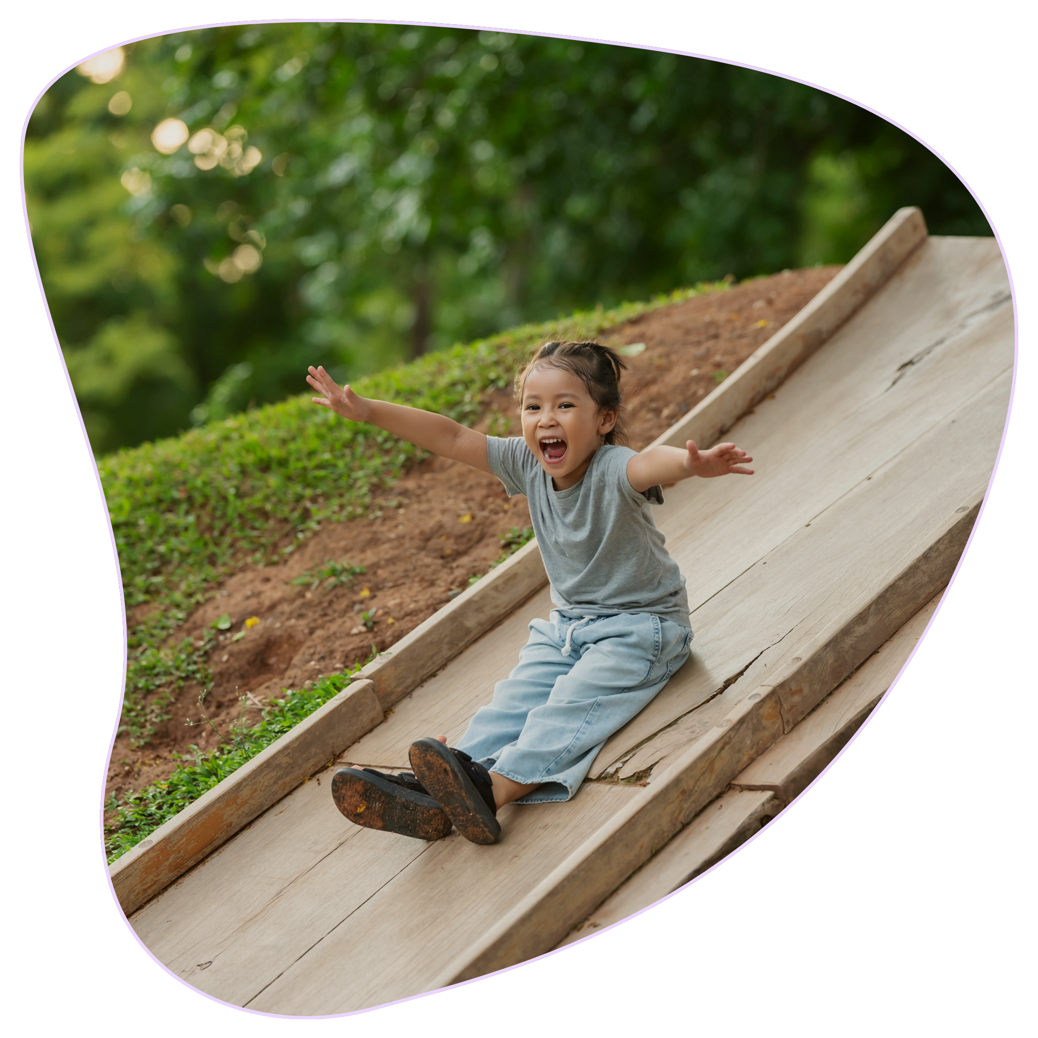 A young girl with braided hair, wearing a gray T-shirt and blue jeans, enjoying a ride on a wooden slide outdoors surrounded by green trees and sunlight because at Strong Roots OT in south NJ and Philadelphia, we provide OT for sensory processing.