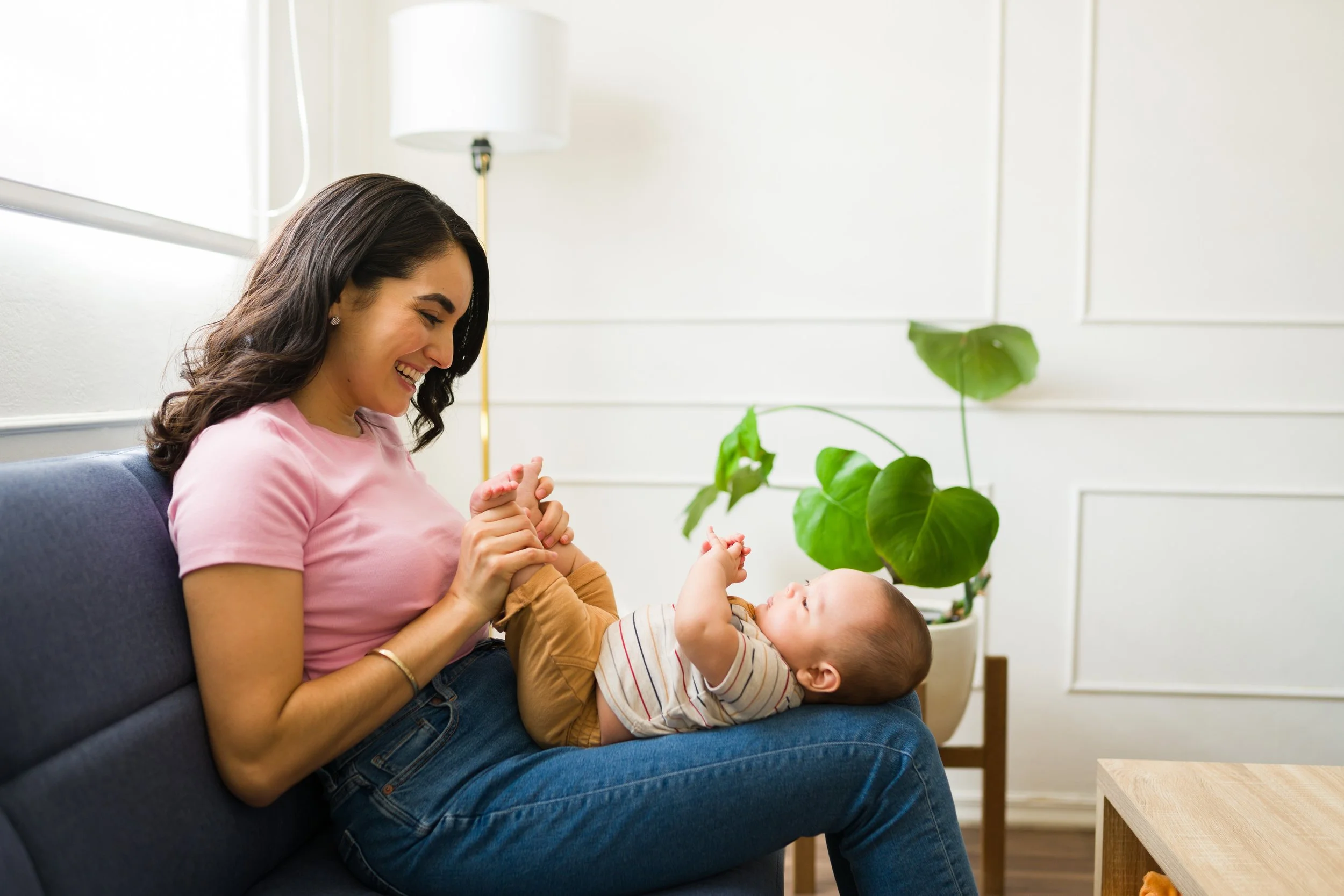 A woman and a baby playing together on a blue sofa in a bright living room with white walls and a large green plant representing Strong Roots OT and the mom and baby bond that we focus on strengthening through occupational therapy in south NJ.