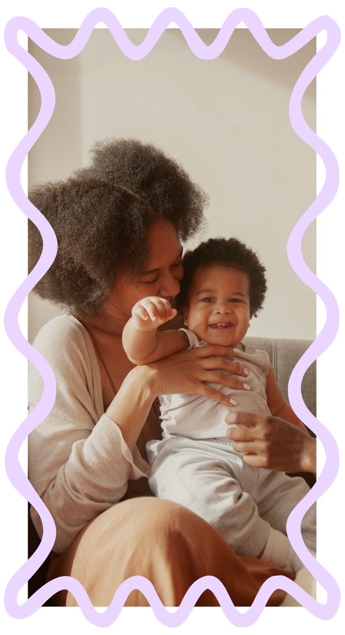 A woman with curly hair holding a smiling toddler with curly hair and light-colored clothing, sitting on her lap in a cozy home setting to represent Strong Roots OT where we provide maternal health & pediatric occupational therapy in south NJ.