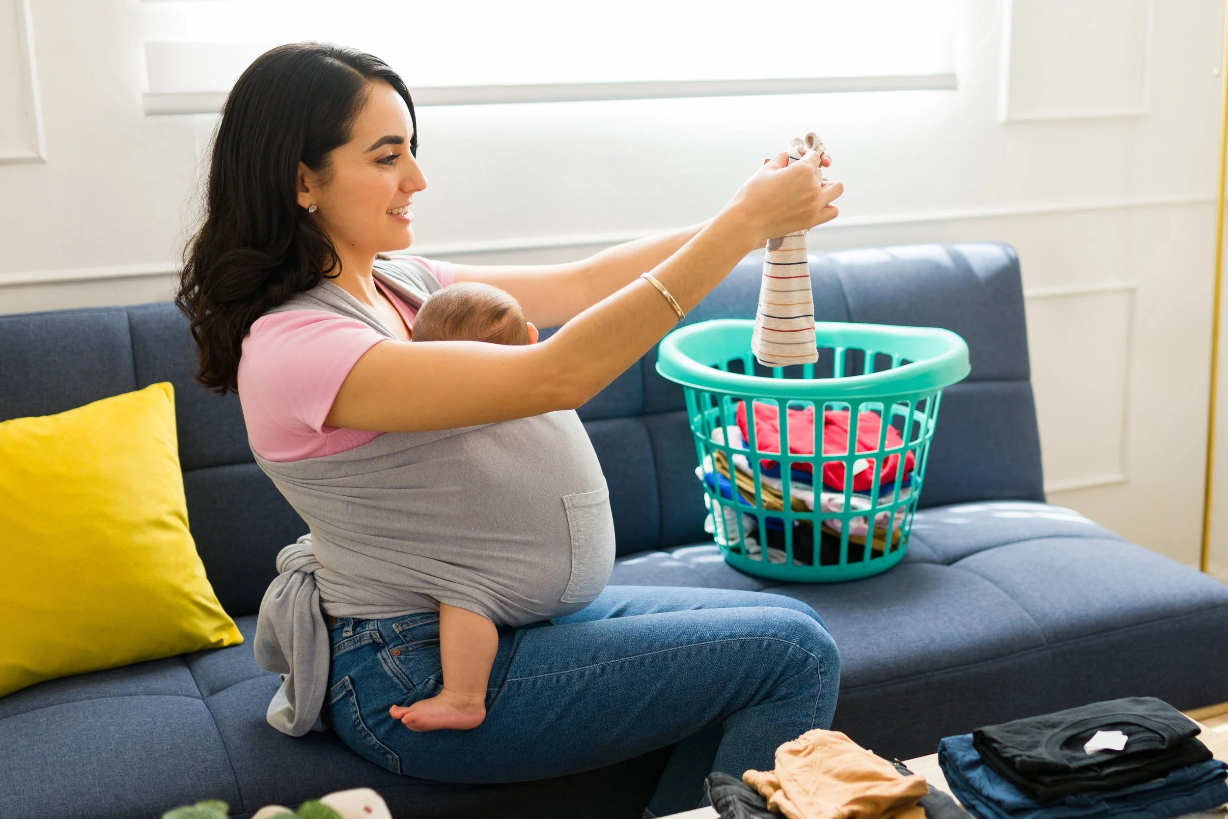 A woman with dark hair, wearing a pink shirt and gray cardigan, is smiling while holding up a striped sock. She is sitting on a blue couch with a yellow pillow. A baby is leaning against her, partially hidden behind her arm for Strong Roots OT.
