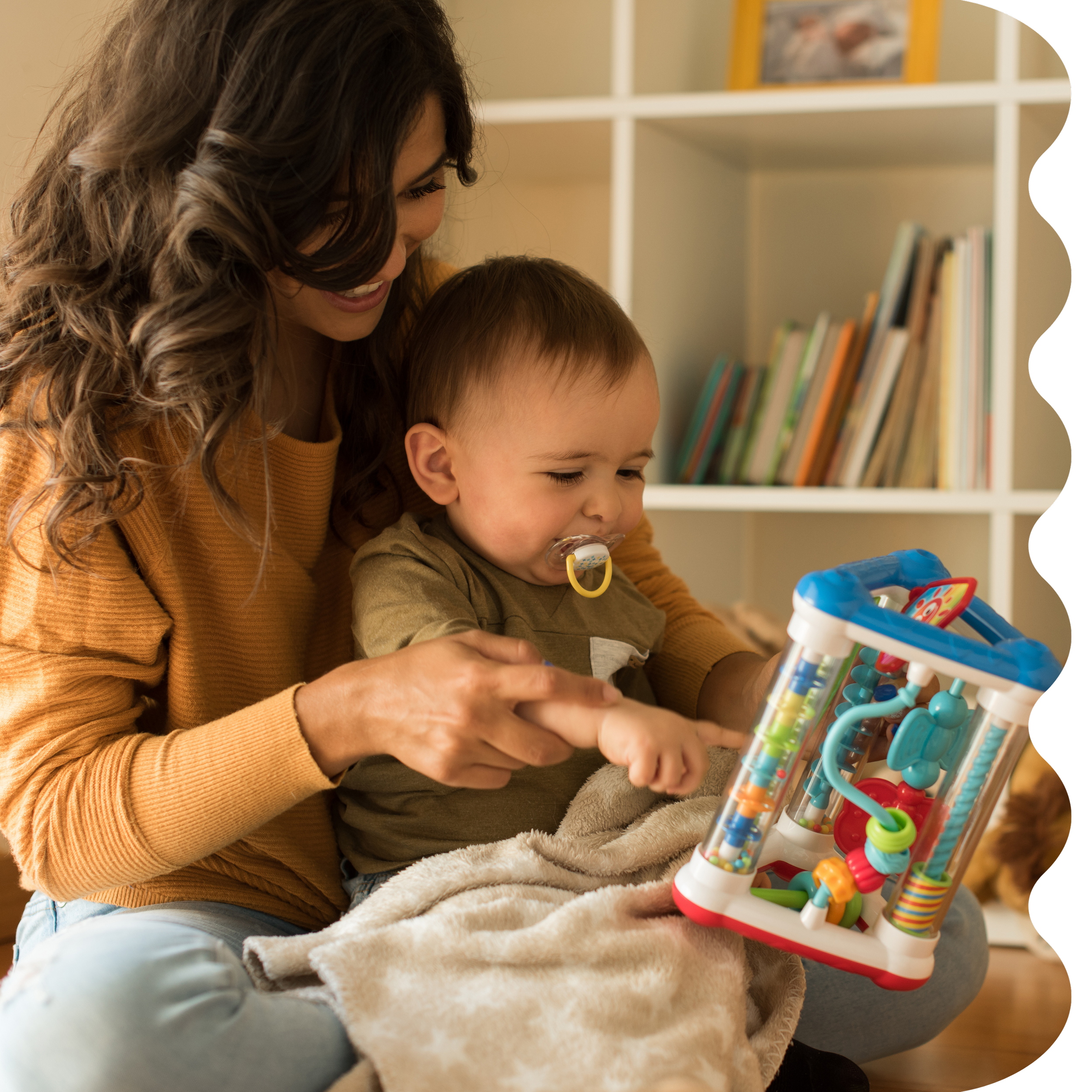 A woman with long, curly brown hair smiling while sitting with a young boy who has a pacifier in his mouth and they are engaging with a colorful tube maze toy. Strong Roots OT provides pediatric OT for older babies and toddlers for fine motor skills.