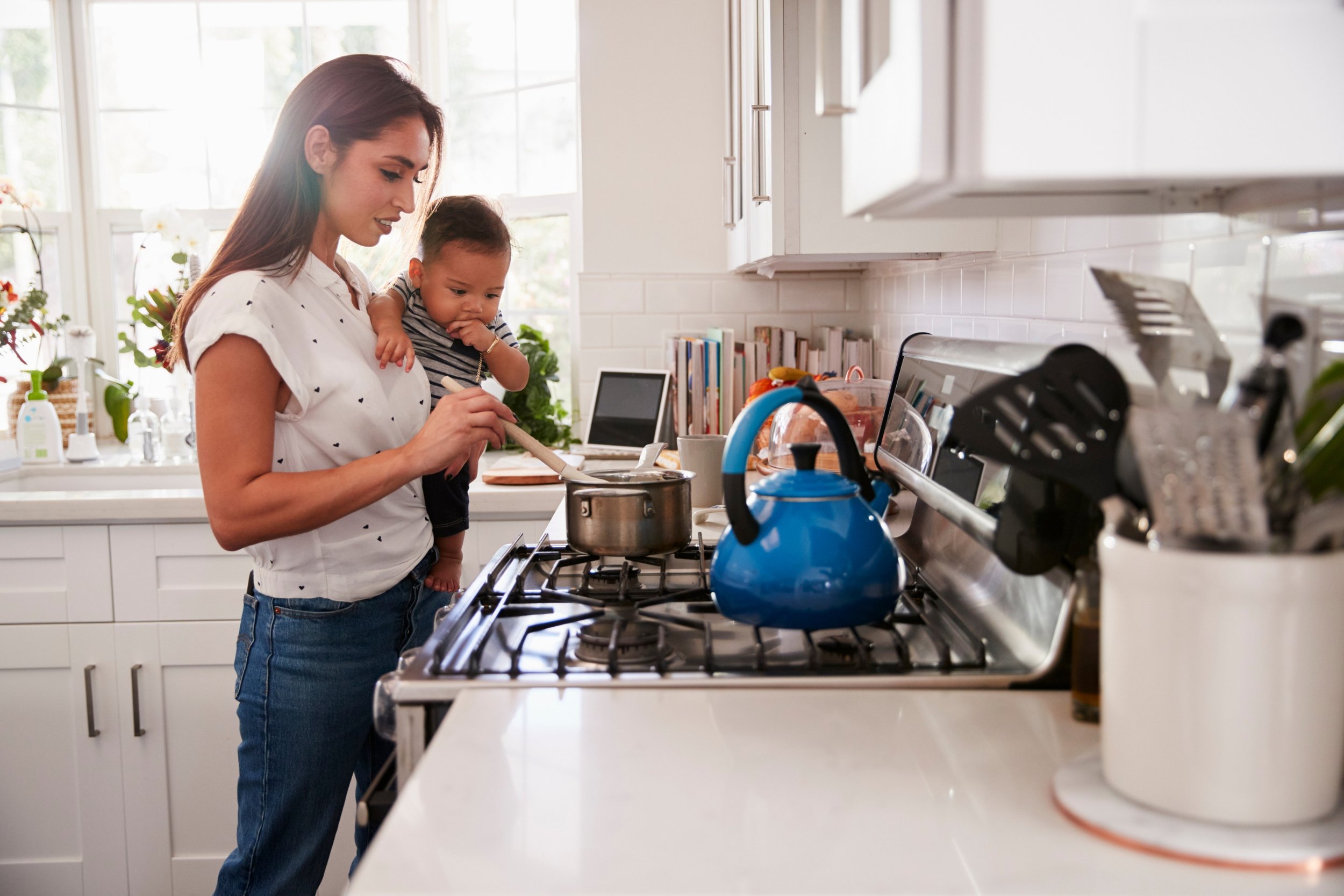 A woman is cooking in a bright, modern kitchen with a child on her hip. The woman is stirring a pot on the stove, and the kitchen counter has various kitchen items, including a blue teapot, books, and plants to represent Strong Roots OT.