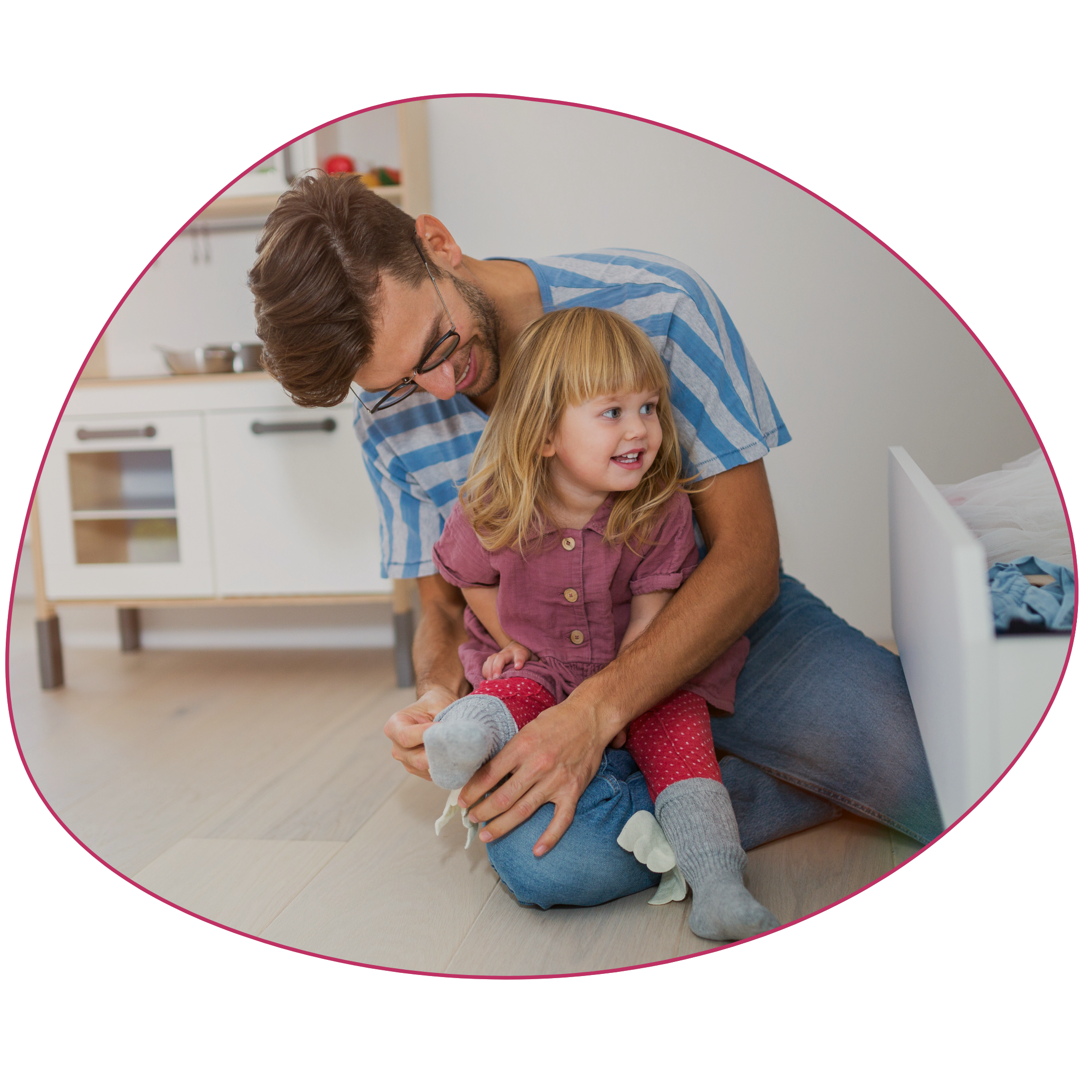 A man with glasses and a beard wearing a blue striped shirt hugging a young girl with long blonde hair in a pink dress, sitting on the floor in a room. Strong Roots OT helps families who are having children struggling with self-help skills.
