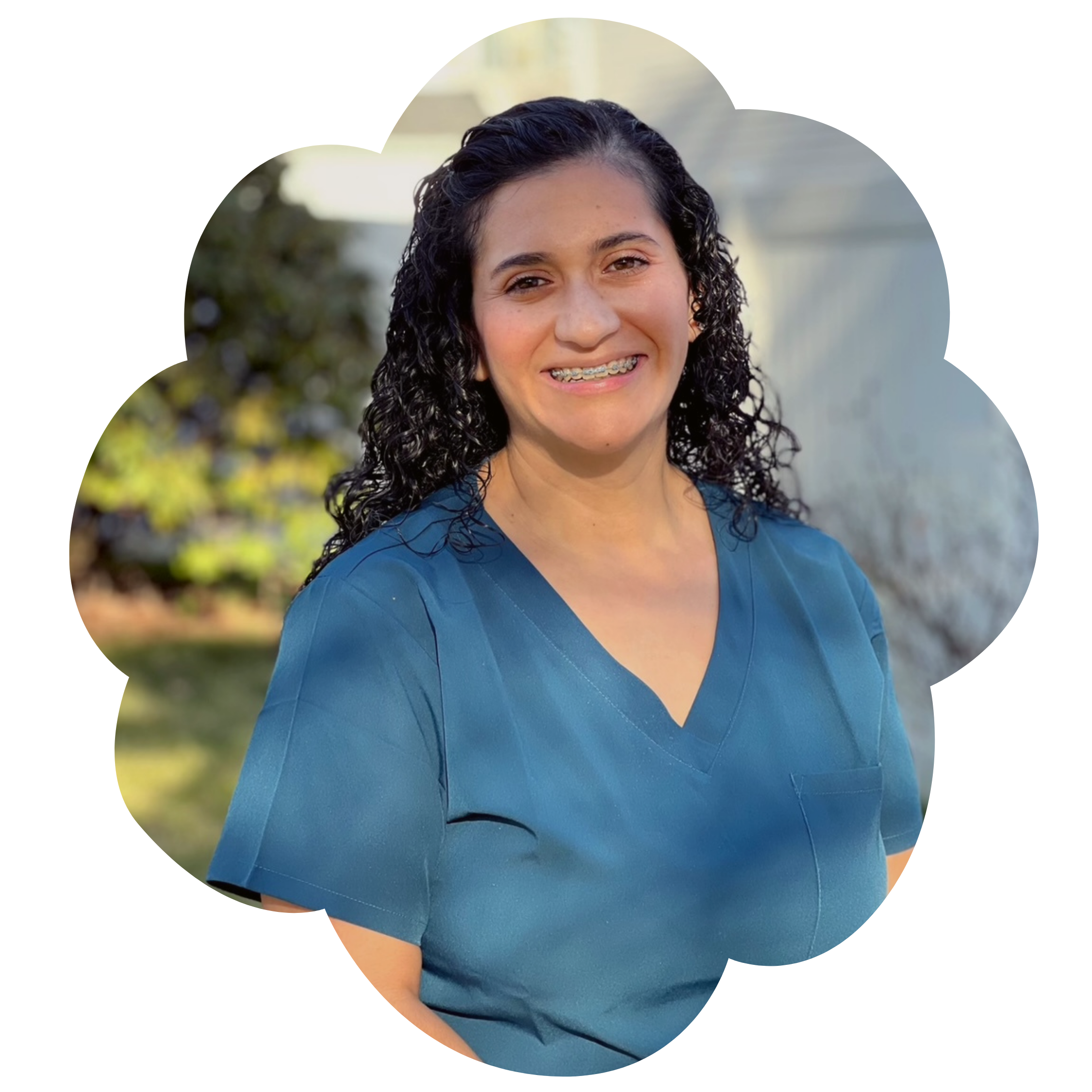A photo of Paula Bradley, the owner of Strong Roots OT and occupational therapist specializing in maternal health and pediatrics. She has curly dark hair, is wearing braces, and is smiling in front of a blurred outdoor background.