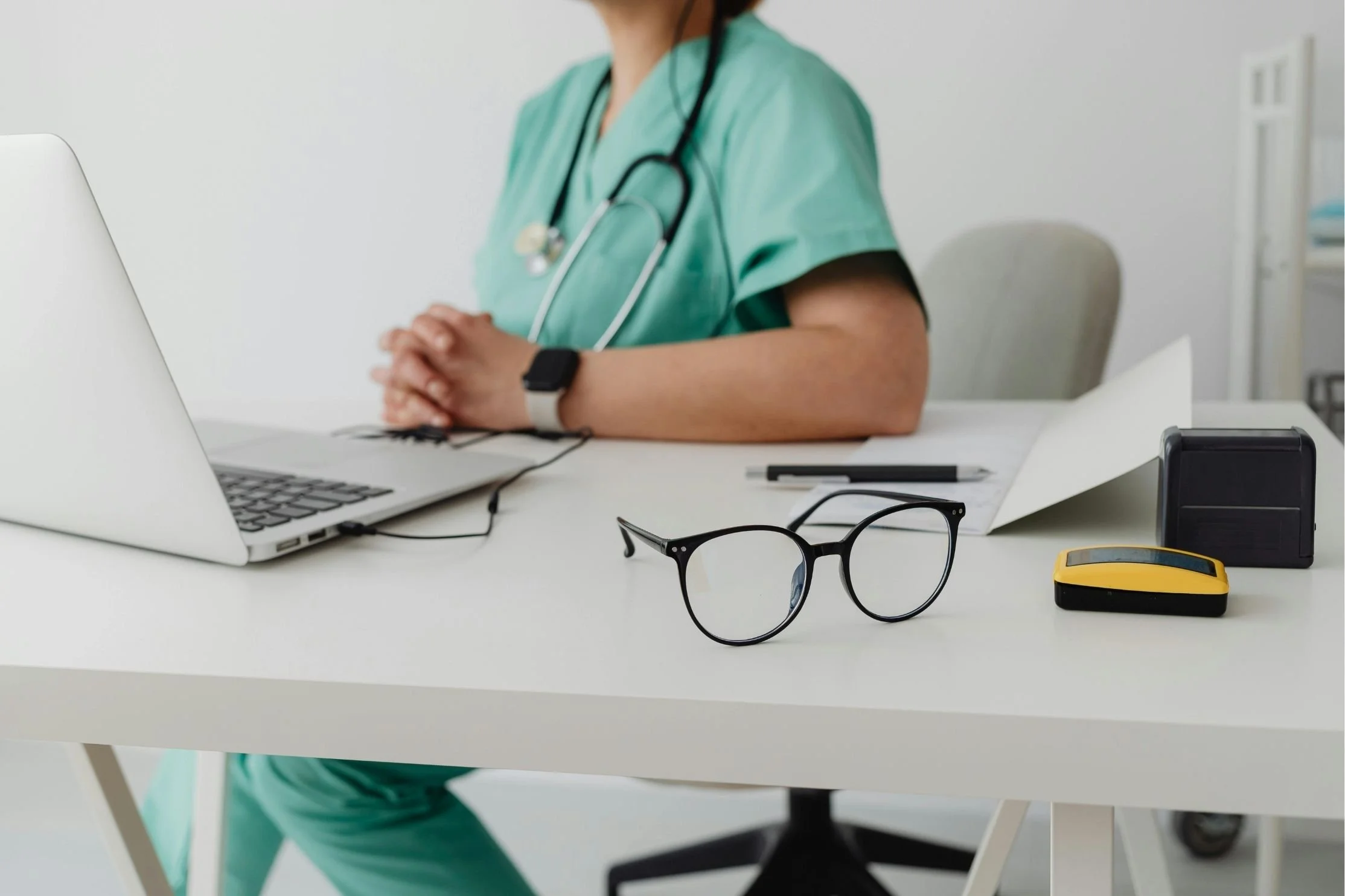 Healthcare professional in medical scrubs sits at a desk with a laptop, documents, and eyeglasses, creating a clean and organized clinical workspace.