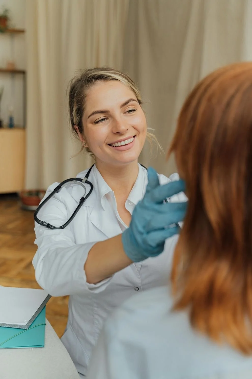 A physician wearing gloves conducts a patient consultation, smiling while performing an examination in a medical office setting.