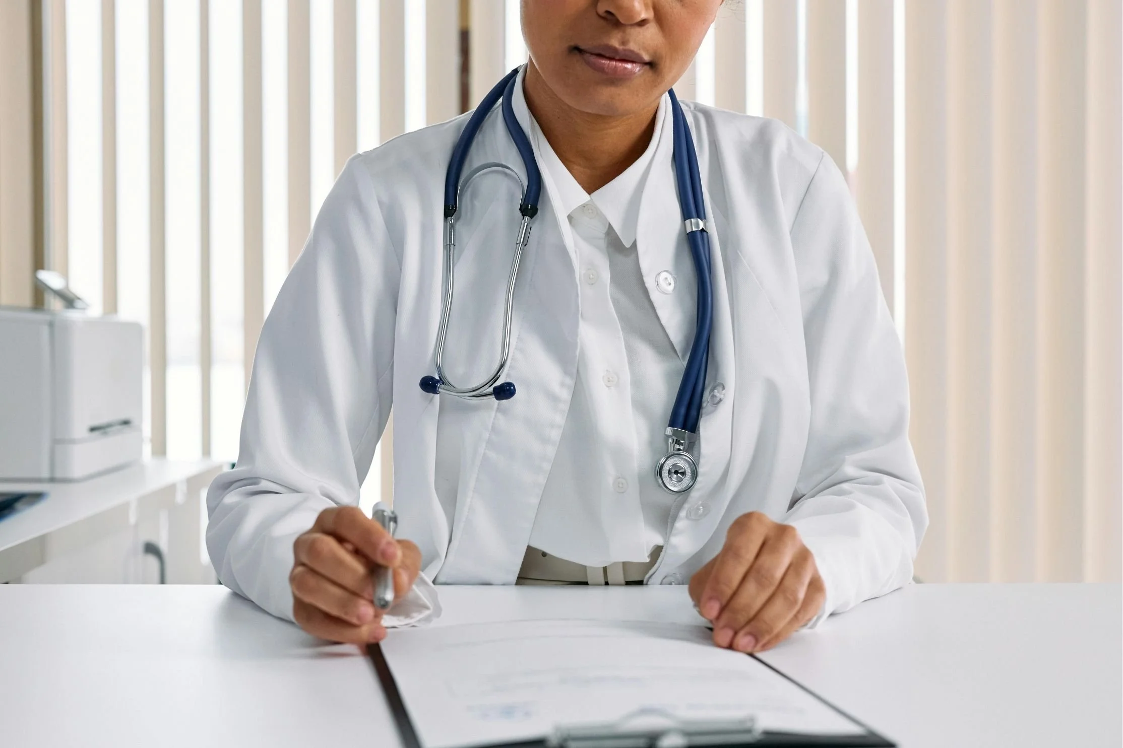 A physician reviews and signs documents at a medical office desk, representing physician coaching and professional development support for healthcare leaders seeking greater effectiveness and career fulfillment.