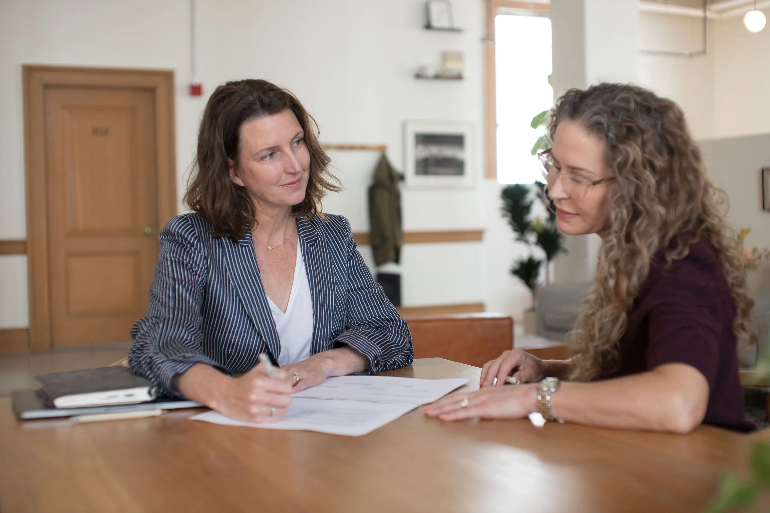 Two women are sitting at a table and talking. One woman has wavy brown hair, wears glasses, and a bun, and is wearing a burgundy top. The other woman has shoulder-length brown hair, wears a striped blazer, and is holding a pen. There are papers on the table between them.