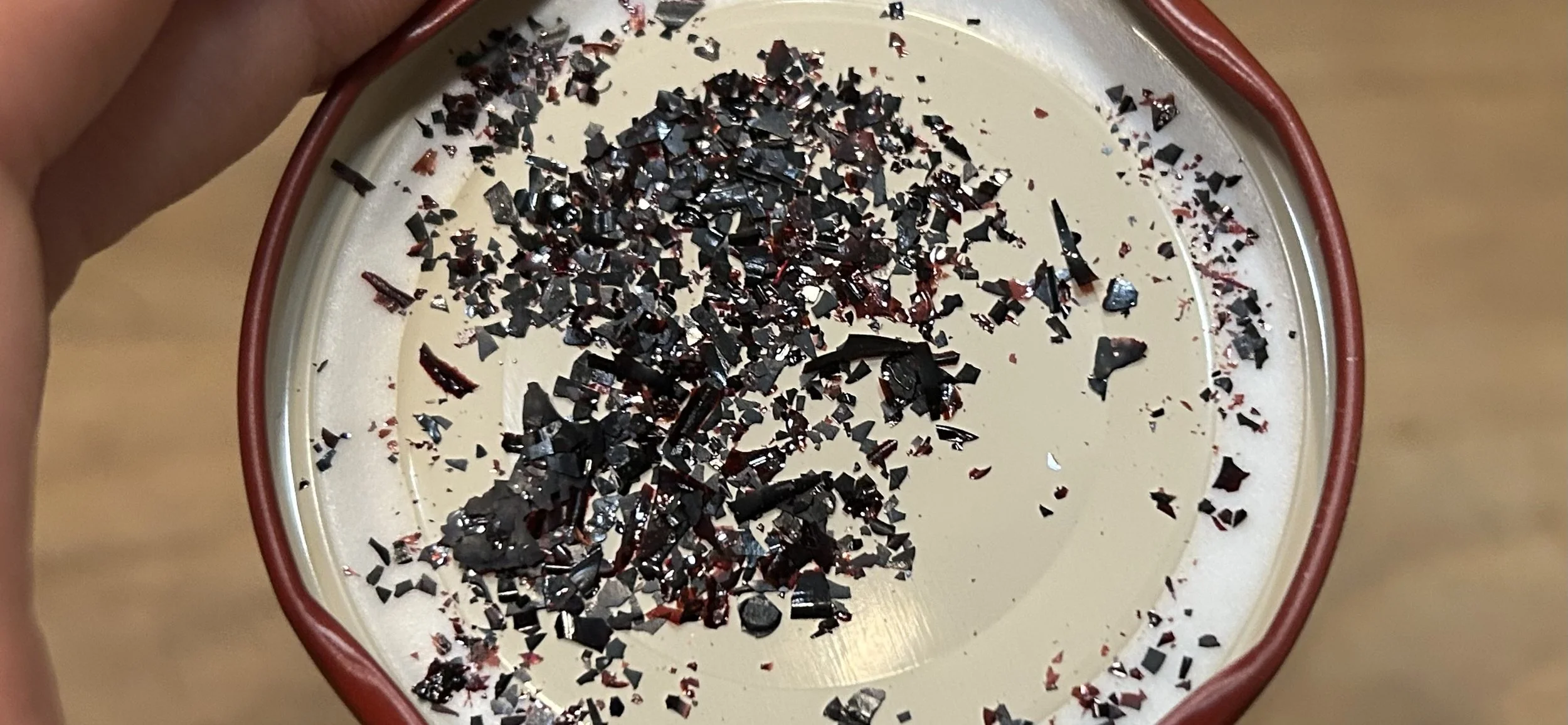 A round ceramic dish with red rim, filled with a creamy white substance and topped with scattered small, shiny black and reddish pieces of crushed chocolate or candy.