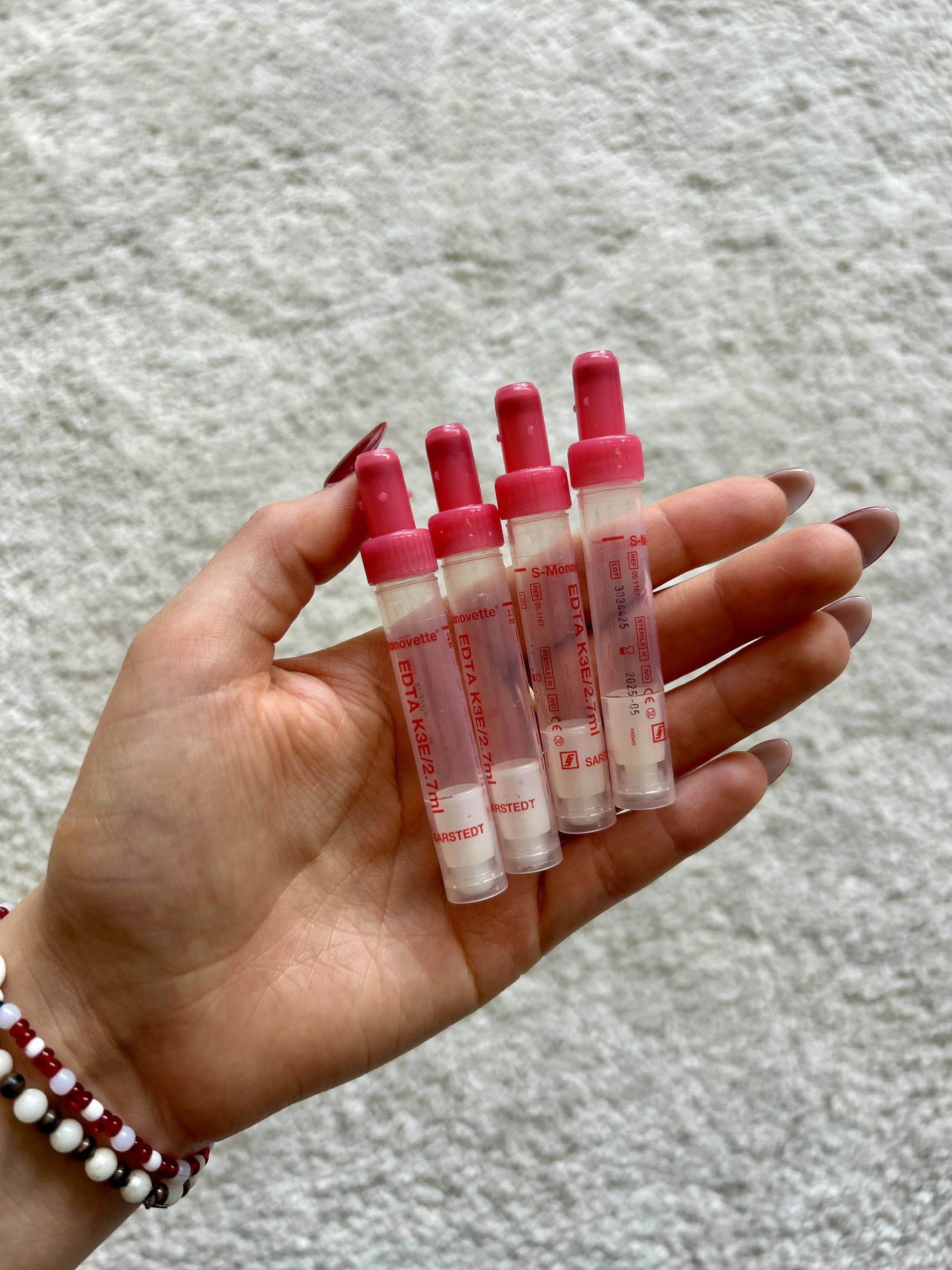 A hand holding six small blood collection tubes with red caps, against a textured beige background. The person is wearing a beaded bracelet with red, white, and black beads.