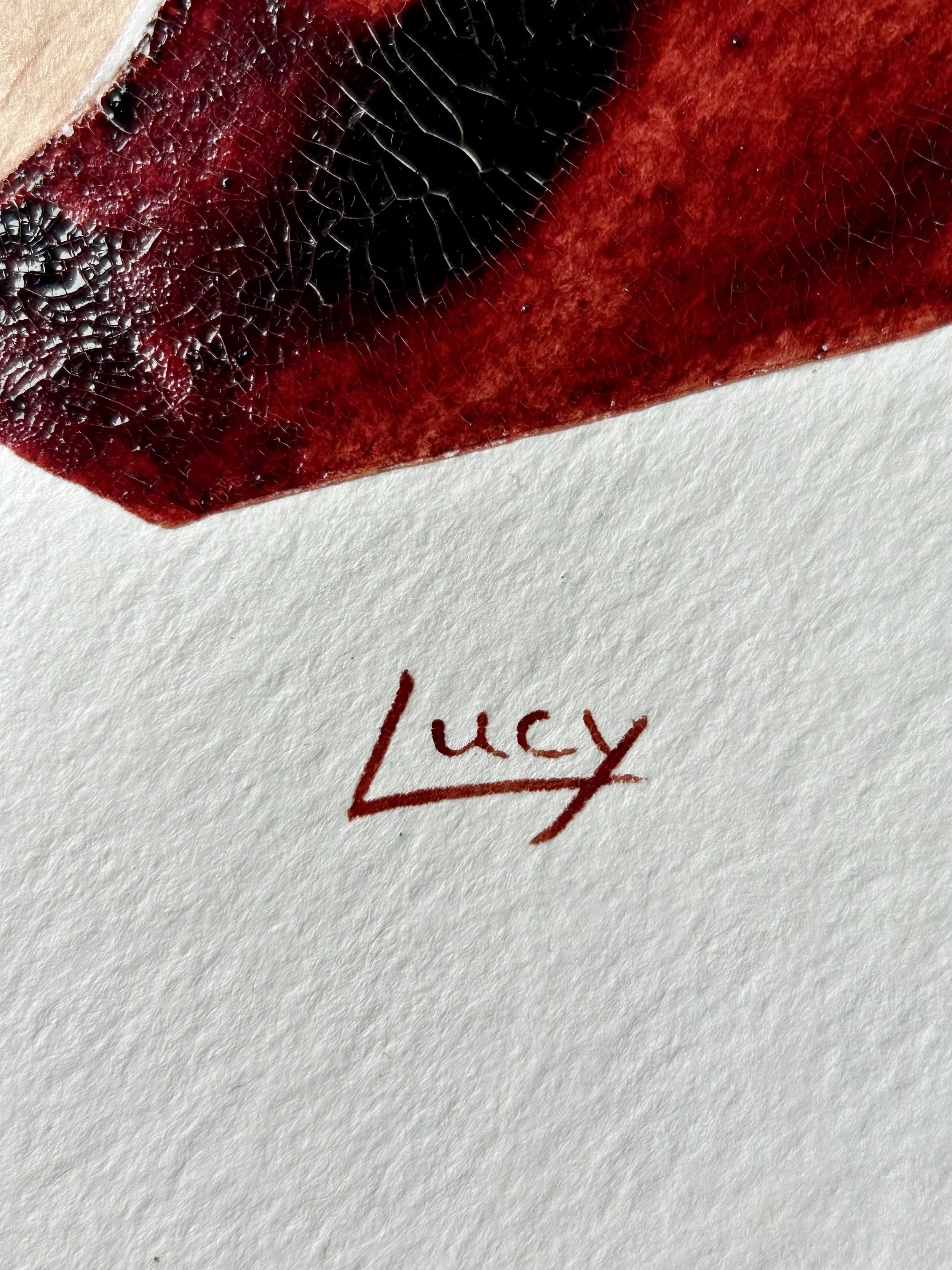 Close-up of a red and black object, possibly a lipstick or cosmetic product, with the word 'Lucy' handwritten in red ink on textured white paper.