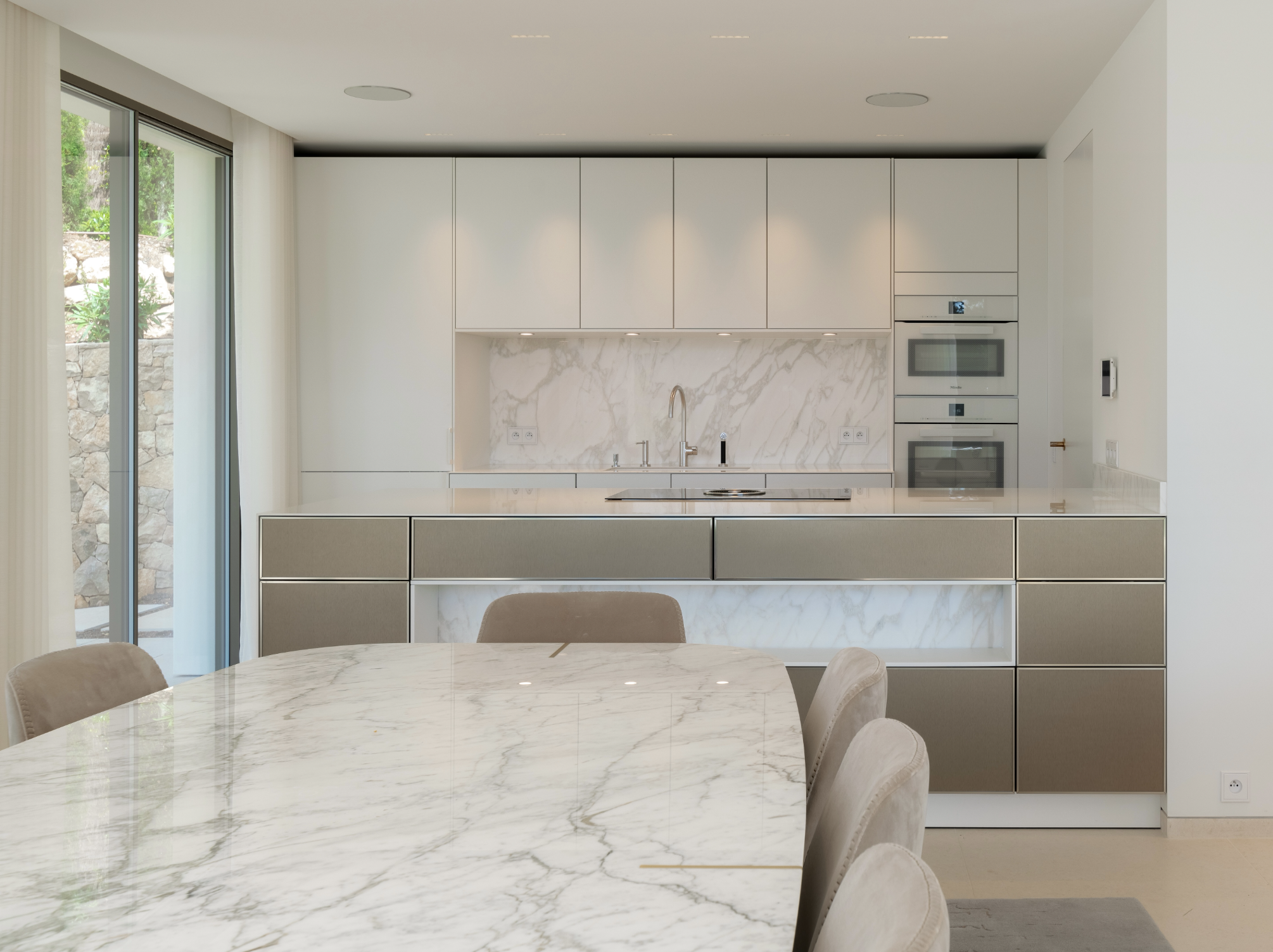 Cuisine moderne avec armoires blanches, un plan de travail en marbre, et un évier en acier inoxydable. Grande table à manger en marbre avec des chaises en velours beige.
