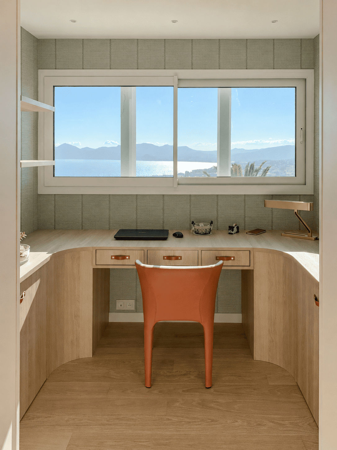 Bureau avec grande fenêtre offrant une vue sur la mer et les montagnes, mobilier en bois, chaise orange, lampe de bureau, ordinateur portable, petits objets de bureau.