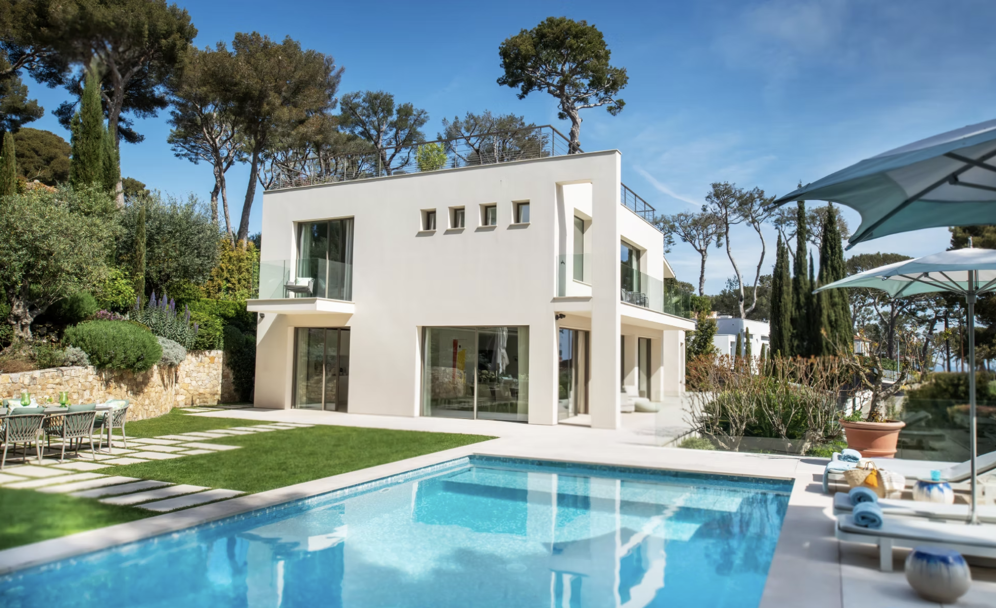 Maison moderne blanche avec grandes fenêtres, piscine à débordement, terrasse, parasols et mobilier d'extérieur, entourée d'arbres et de jardin luxuriant.