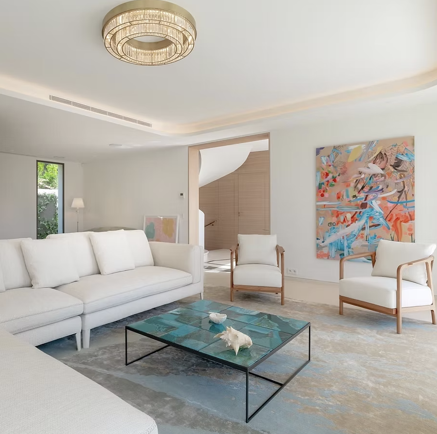 Salon moderne avec canapé blanc, deux fauteuils avec cadre en bois, table basse en verre, tableau abstrait coloré sur le mur et lampe de plafond en cercle.