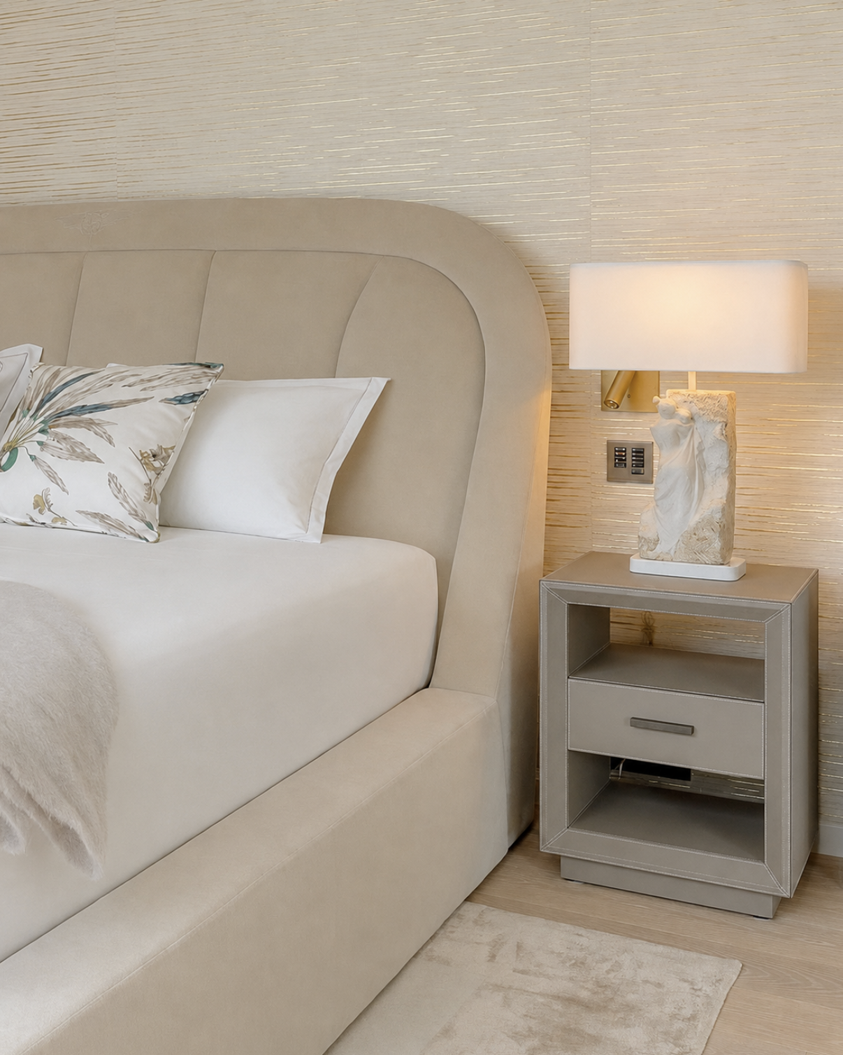 L'intérieur d'une chambre avec un lit beige, une tête de lit rembourrée, et une table de nuit grise avec une lampe de style sculpture en pierre et un abat-jour blanc.