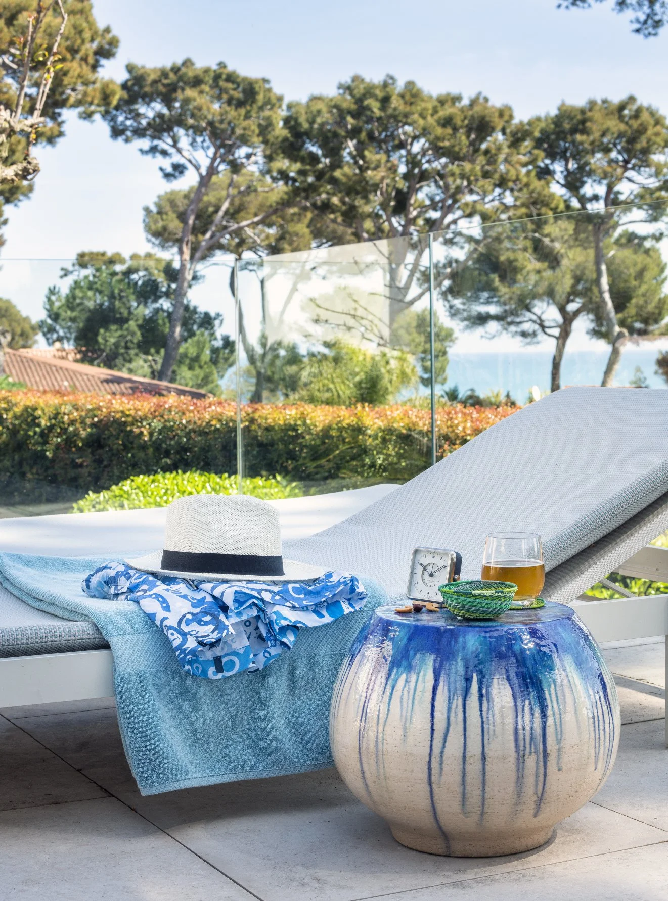 Une chaise longue blanche avec un coussin gris, une serviette bleue, un chapeau blanc avec un ruban noir, un verre de boisson, une horloge, un bol vert et une surface bleue en verre, sur un patio extérieur avec vue sur des arbres et la mer en arrière-plan.