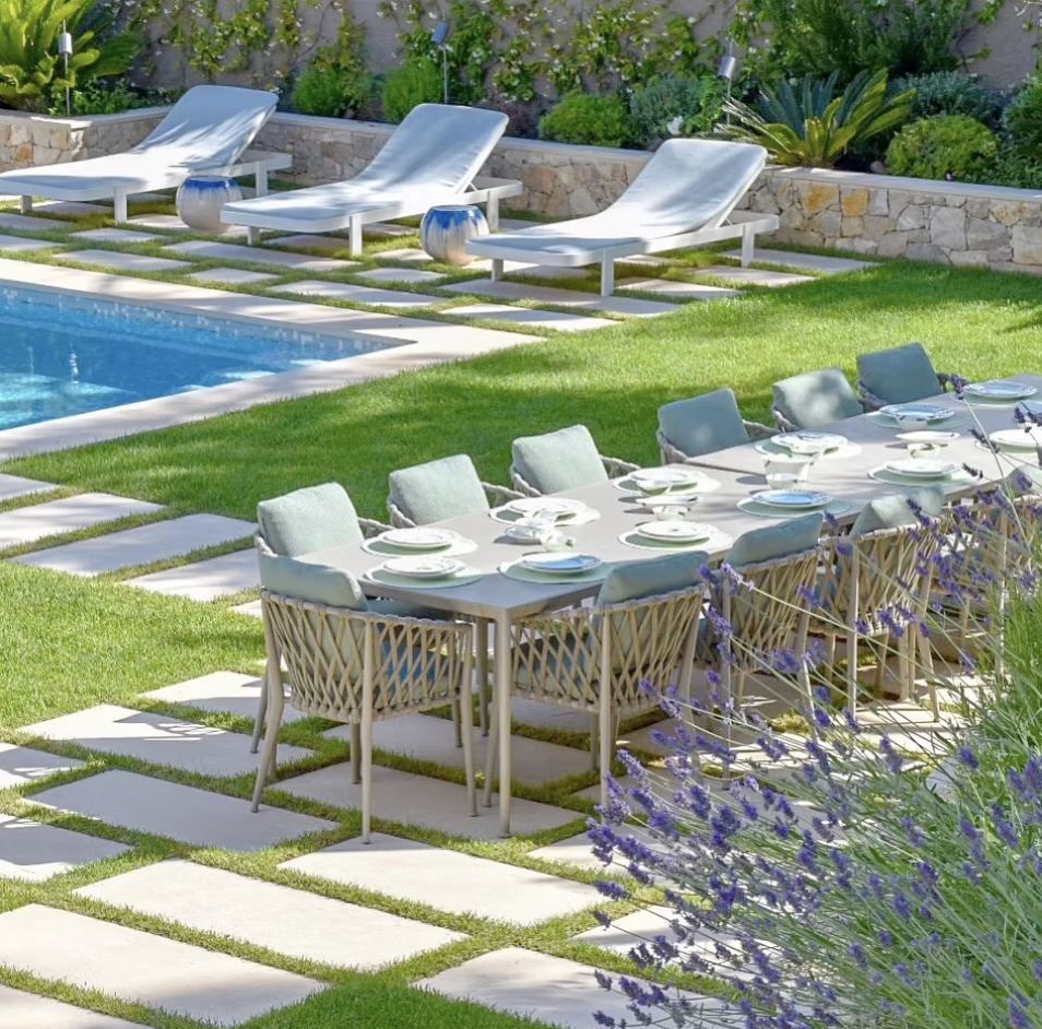 Une table de dîner est préparée avec des assiettes, des verres et des couverts dans un jardin avec une piscine, des chaises longues et des plantes vertes.