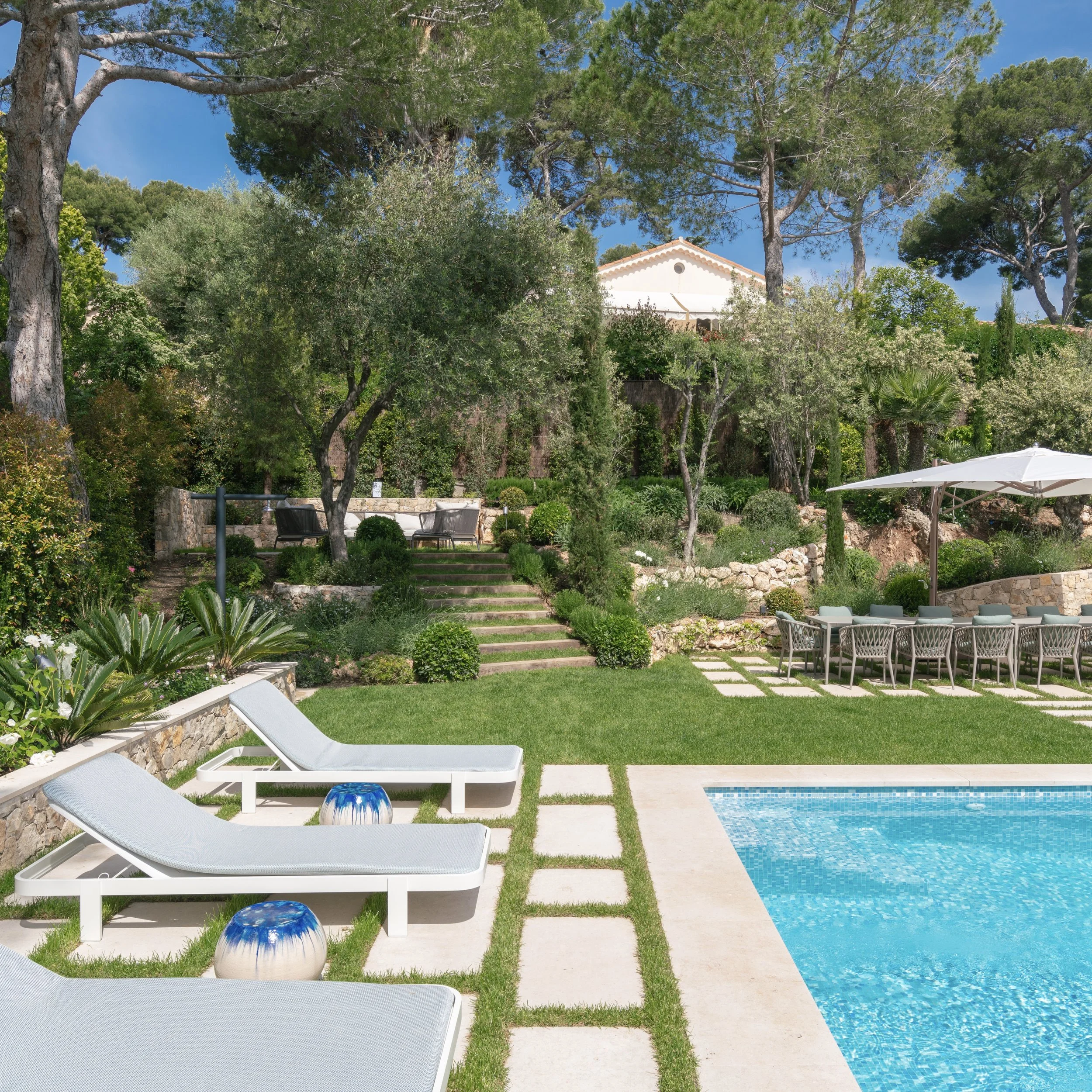 Un jardin avec des chaises longues blanches, un parasol blanc et une piscine. Il y a beaucoup d'arbres et un ciel bleu.