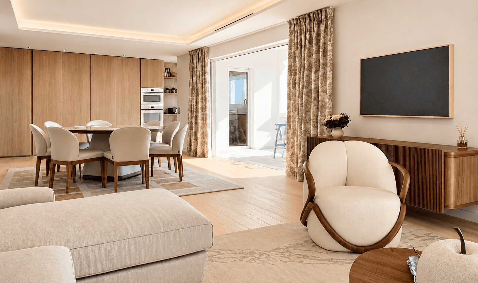 Salon avec canapé beige, fauteuil blanc, table basse en bois, mur avec grand tableau noir, porte-fenêtre donnant sur un balcon lumineux, cuisine en arrière-plan, décor moderne avec meubles en bois clair et plantes