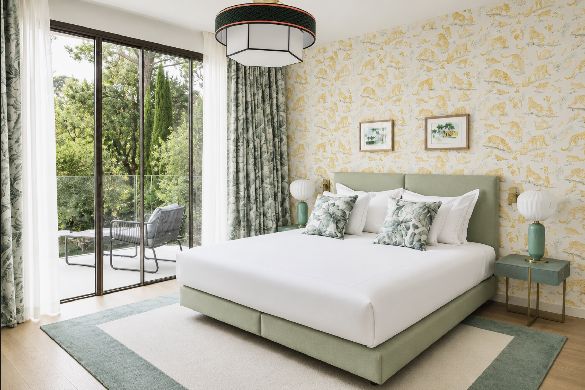 Chambre avec un grand lit à tête verte, rideaux à motifs de feuilles, deux lampes de chevet, une table avec une lampe en forme de boule, deux tableaux avec des arbres, un balcon avec des chaises, murs avec papier peinte de lions et autres animaux.
