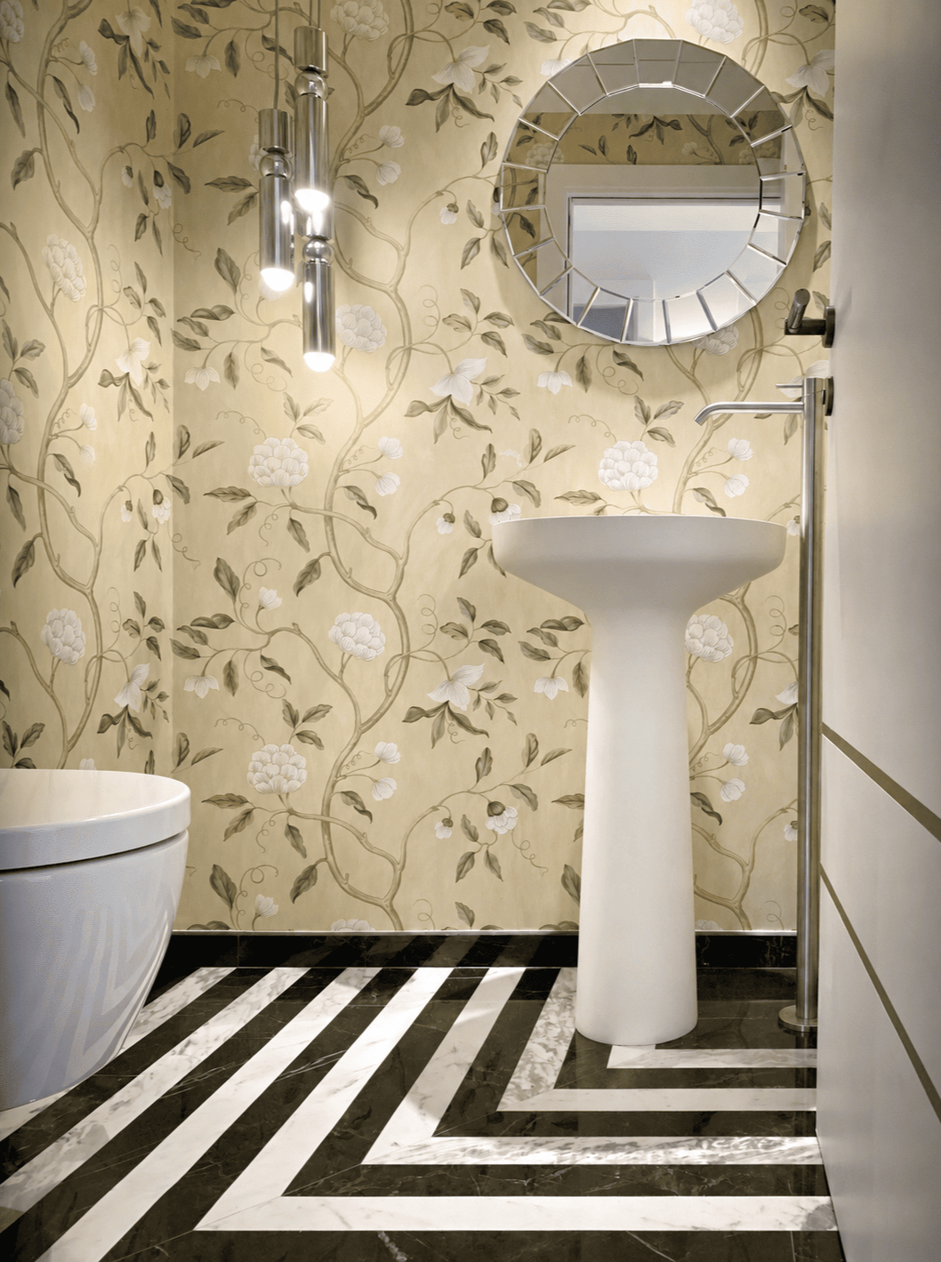 Salle de bain élégante avec papier peint floral beige, miroir rond, lavabo sur pied blanc, toilette, et éclairage suspendu moderne.