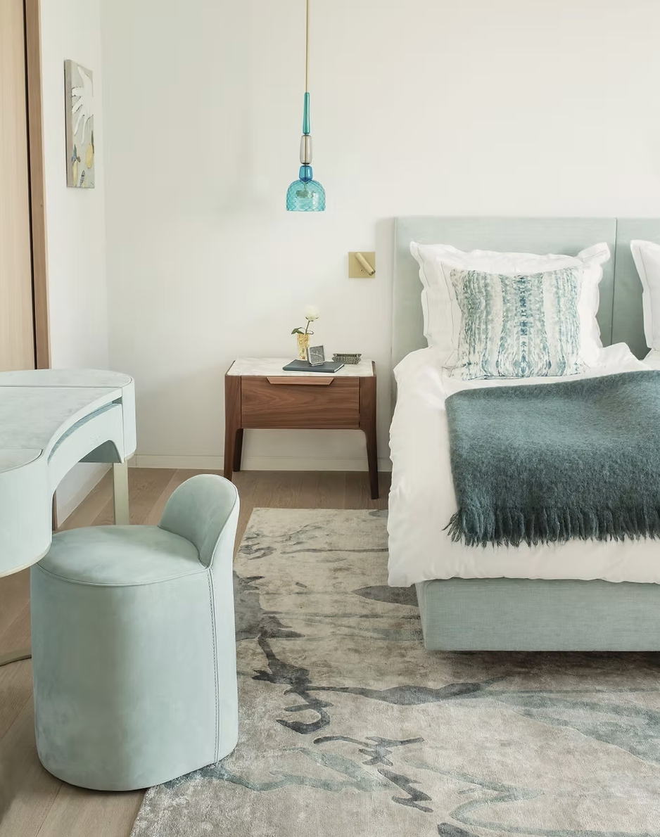 Chambre avec lit avec oreillers et une couverture, une table de chevet en bois avec une petite plante et une lampe suspendue bleu, une chaise verte et une table ronde en blanc, mur blanc avec une peinture murale