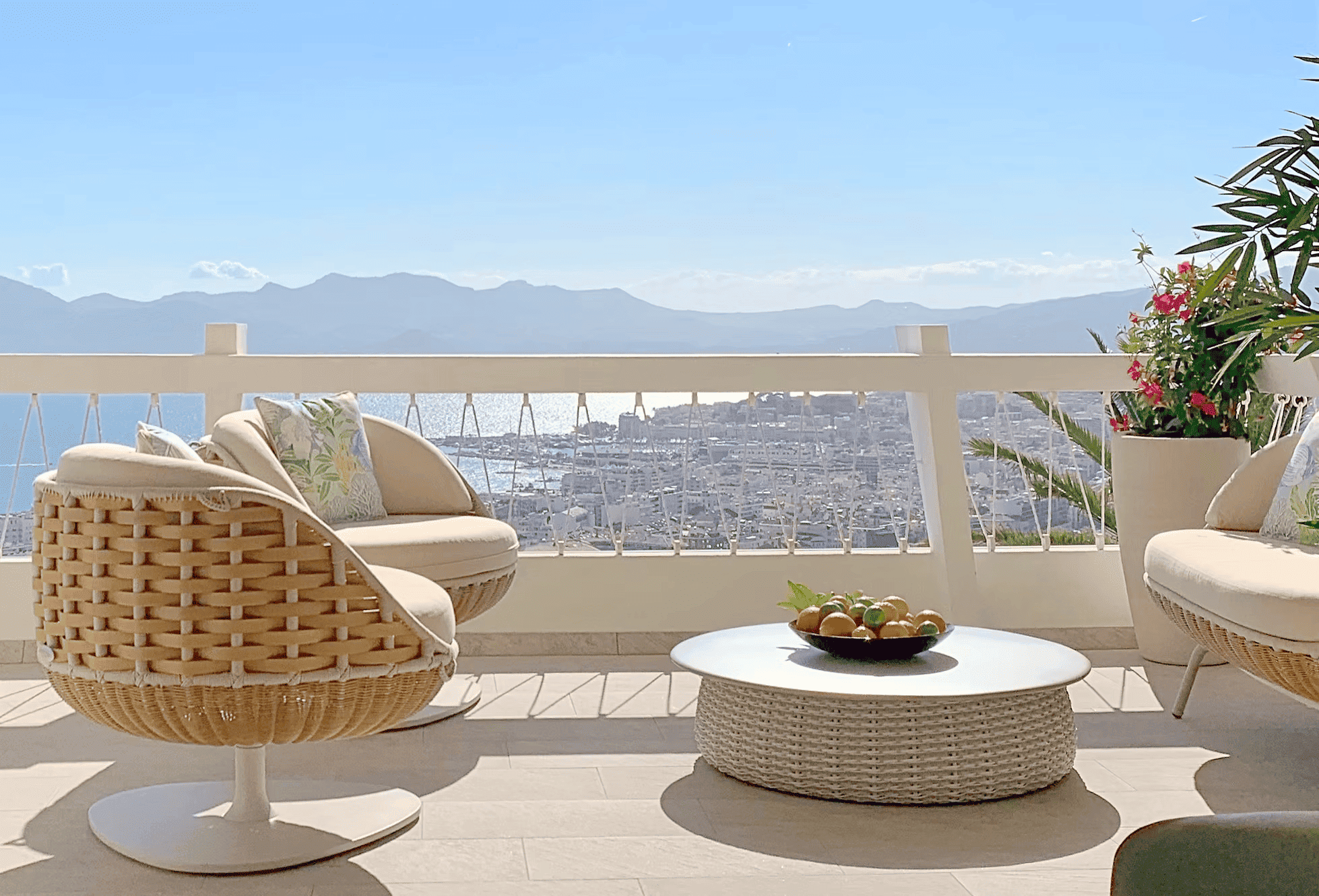Terrasse extérieure avec mobilier en rotin, vue sur la mer et les montagnes au loin, avec un ciel clair, une pièce verte avec des fleurs roses, et une table basse ronde avec un bol de fruits.