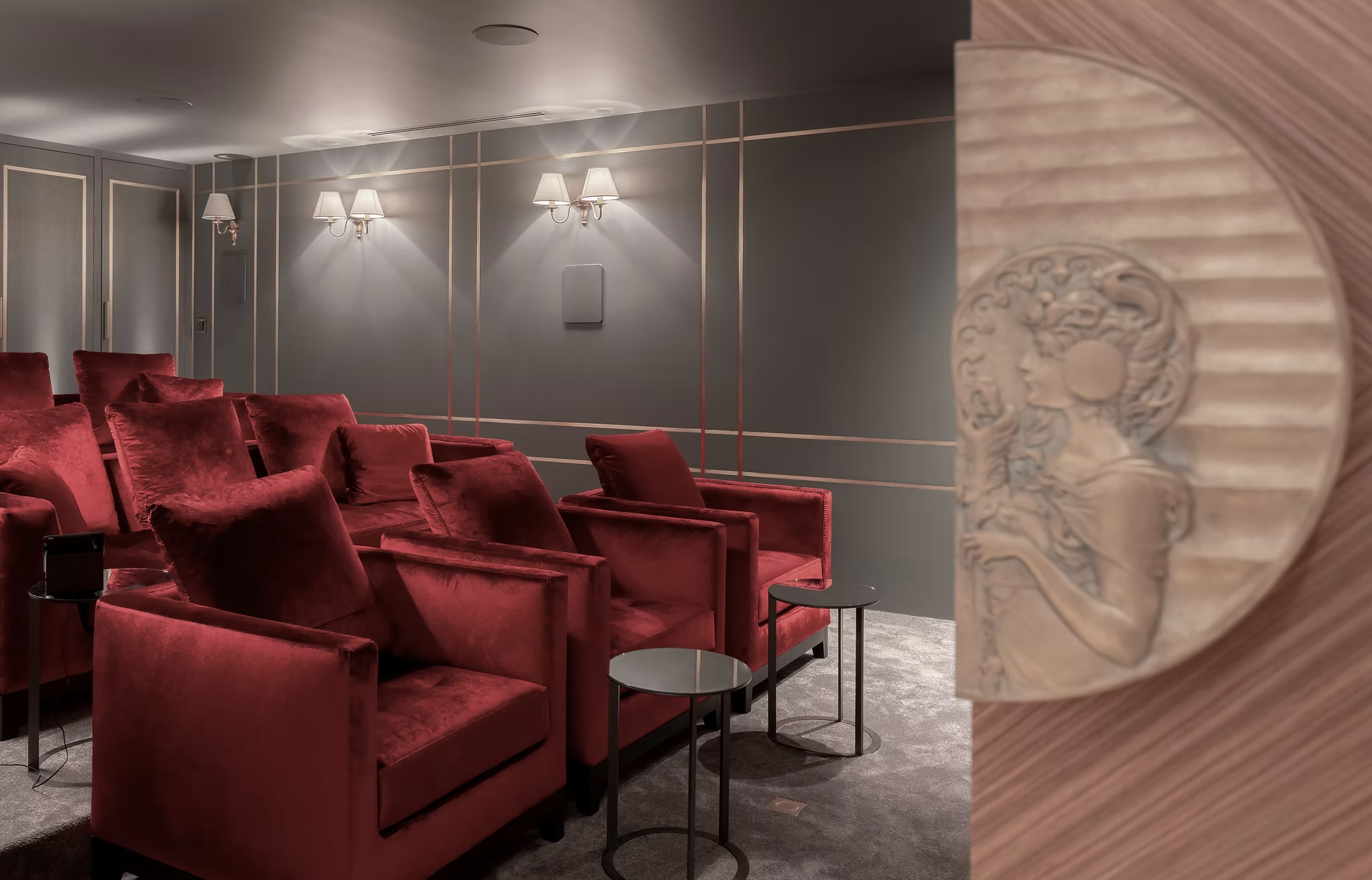 Salle de cinéma avec des fauteuils rouges, murs gris avec moulures, appliques murales avec abat-jour, et pièce en bois sculpté visible sur le côté droit.