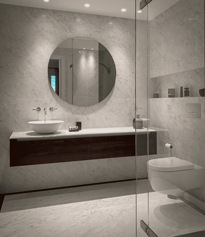 Salle de bain moderne avec murs en marbre, miroir rond, évier sur un meuble en bois, décor minimaliste avec quelques objets et vasques sur une étagère, toilettes modernes, porte en verre.