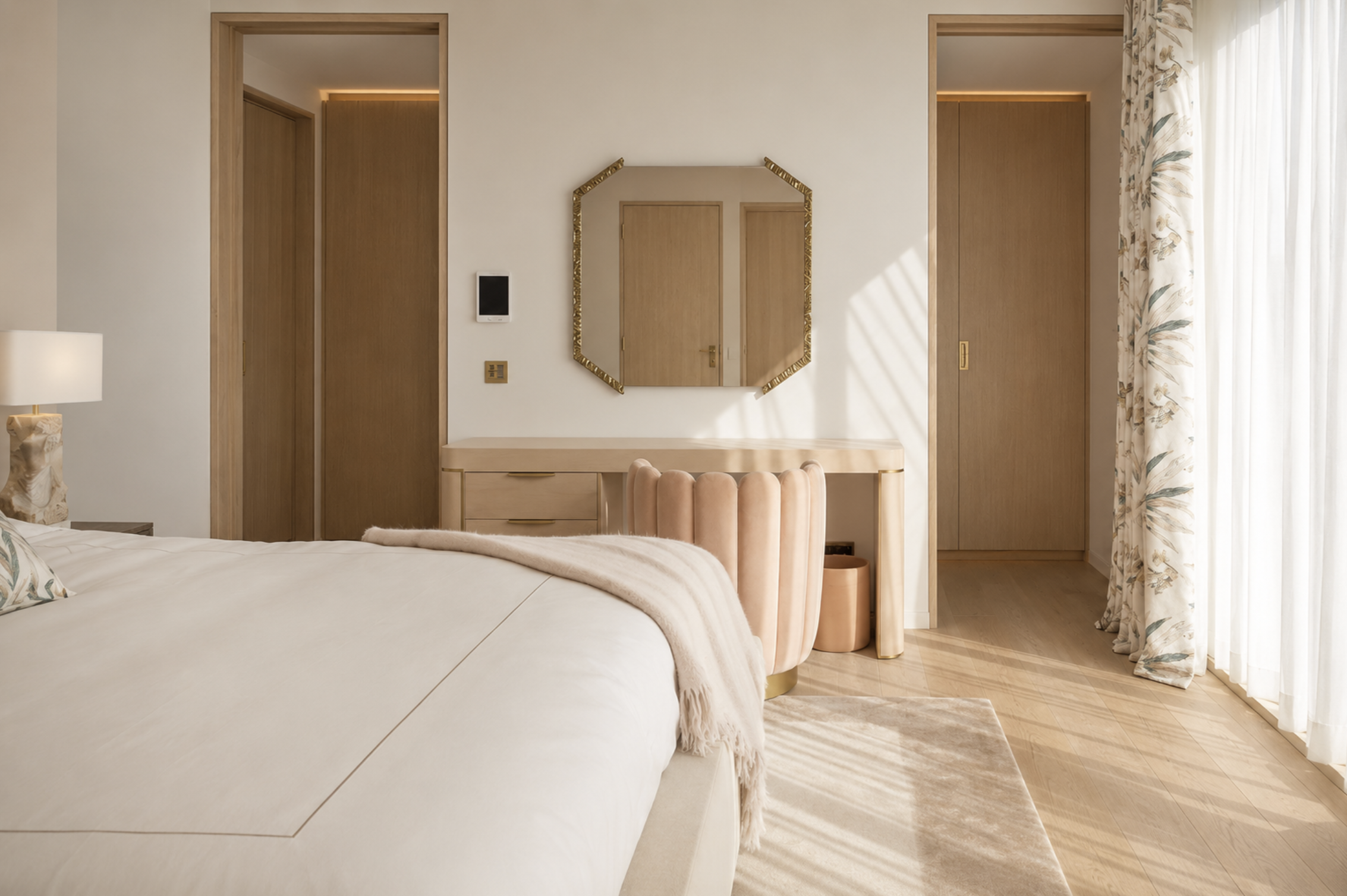 Un coin d'une chambre moderne avec un lit blanc, un miroir avec cadre doré, une commode en bois clair, une chaise en velours beige, une table de chevet, une lampe, et de grandes fenêtres avec des rideaux floraux laissant entrer la lumière naturelle.