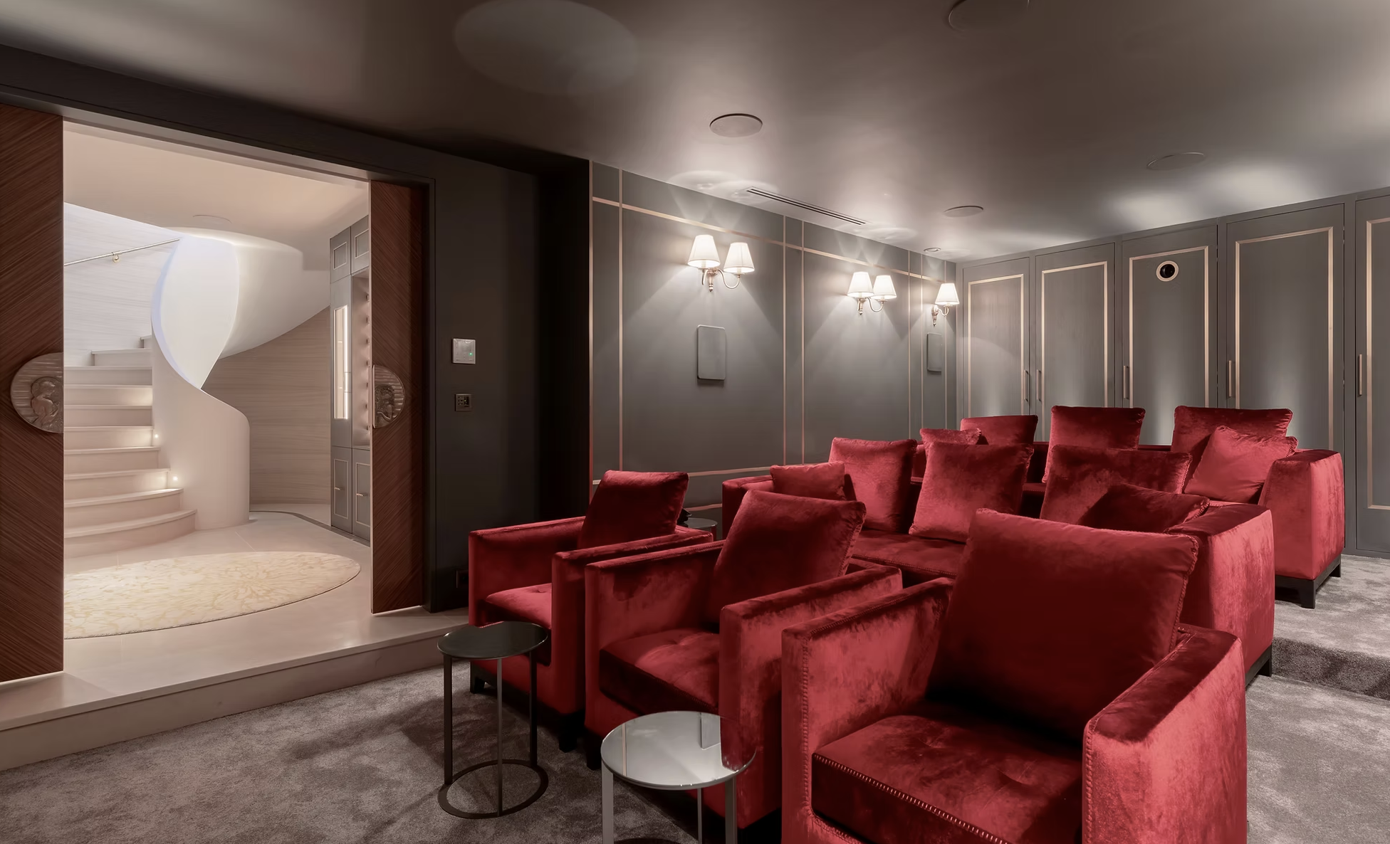 Salle de séjour avec canapé en velours rouge, fauteuils, petites tables, murs gris avec moulures, appliques murales, lumière douce, escalier en spirale blanc visible par la porte cochère.