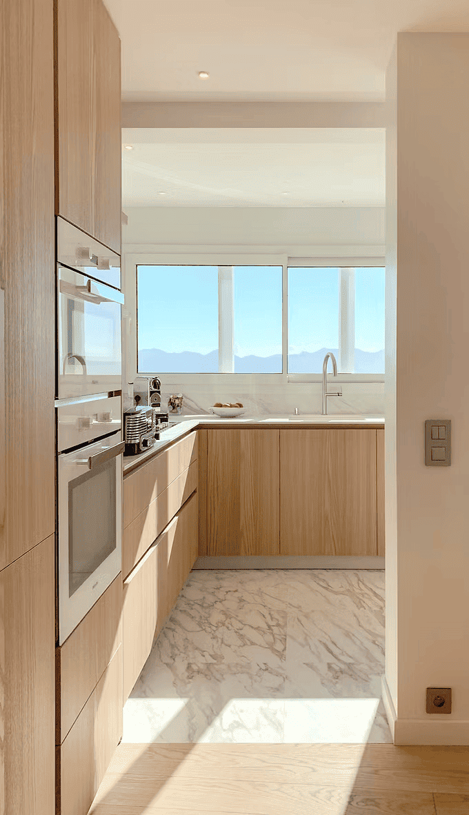 Cuisine moderne avec armoires en bois clair, comptoir, appareils électroménagers en acier inoxydable, grande fenêtre offrant une vue sur des montagnes et un ciel bleu.