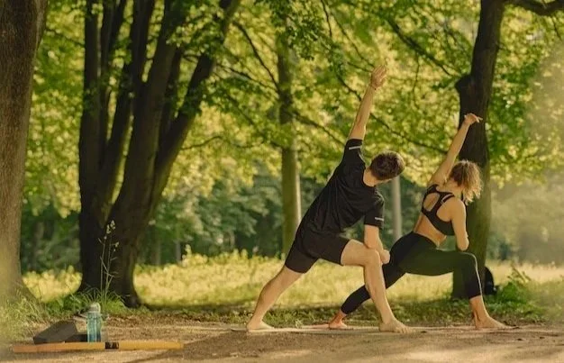 A man and woman practicing yoga outdoors in a wooded park, performing a side angle pose together.