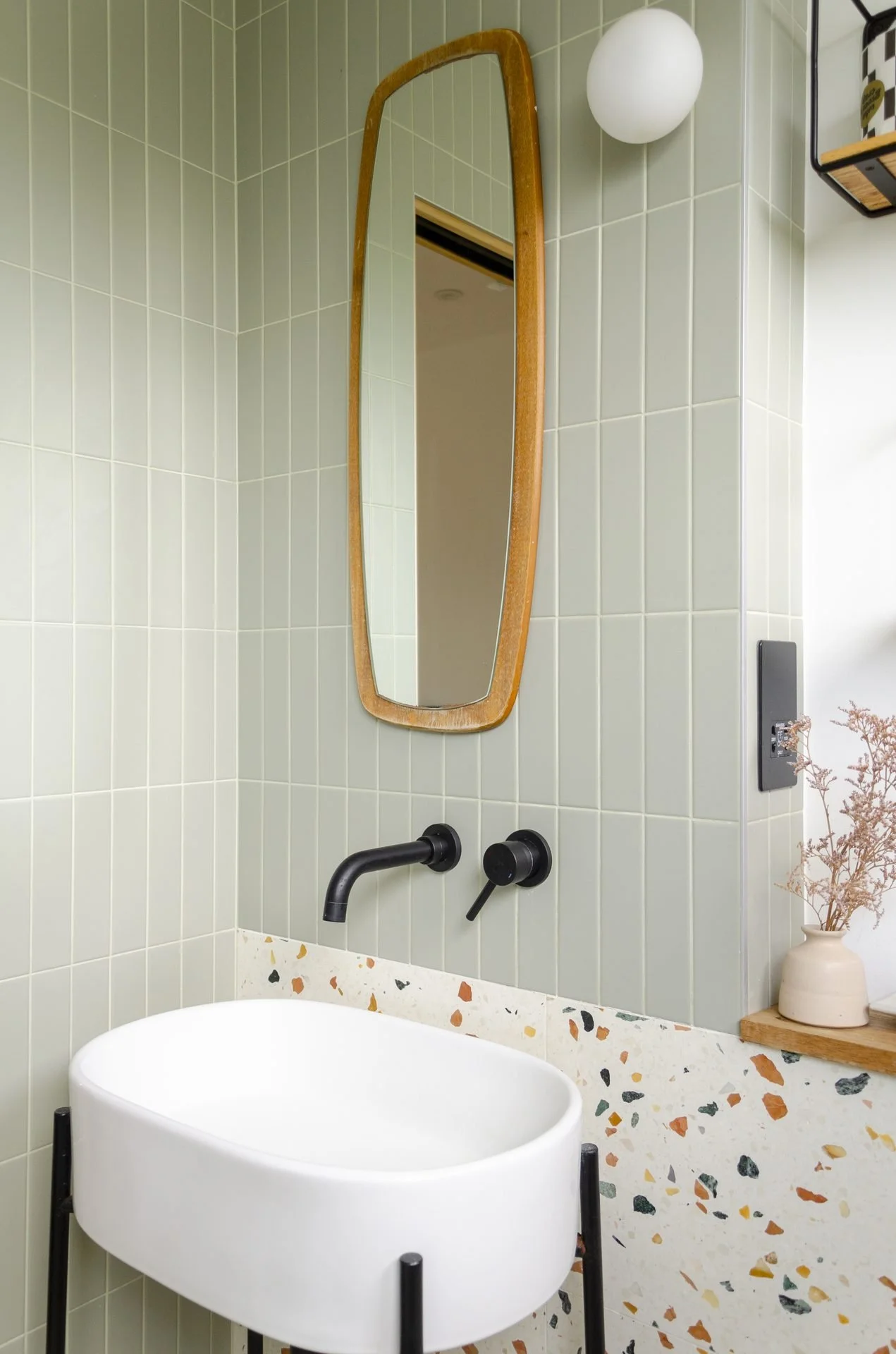 Bathroom with a terrazzo counter, a matte black faucet, a small white oval bathtub, a rectangular mirror with a rounded wooden frame, a white spherical wall light, a small potted plant, and a green tiled wall.