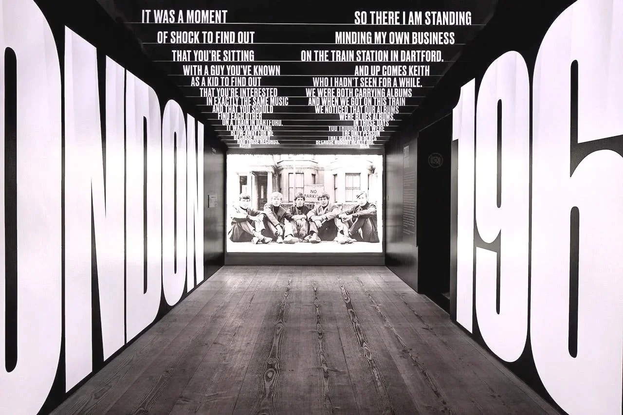 Black and white hallway with large text on the walls spelling out "INDUSTRY" and "MUSIC" and musical lyrics painted on the ceiling. At the end of the hallway, there is a window showing a black and white photo of five young men sitting on a ledge outside a building.