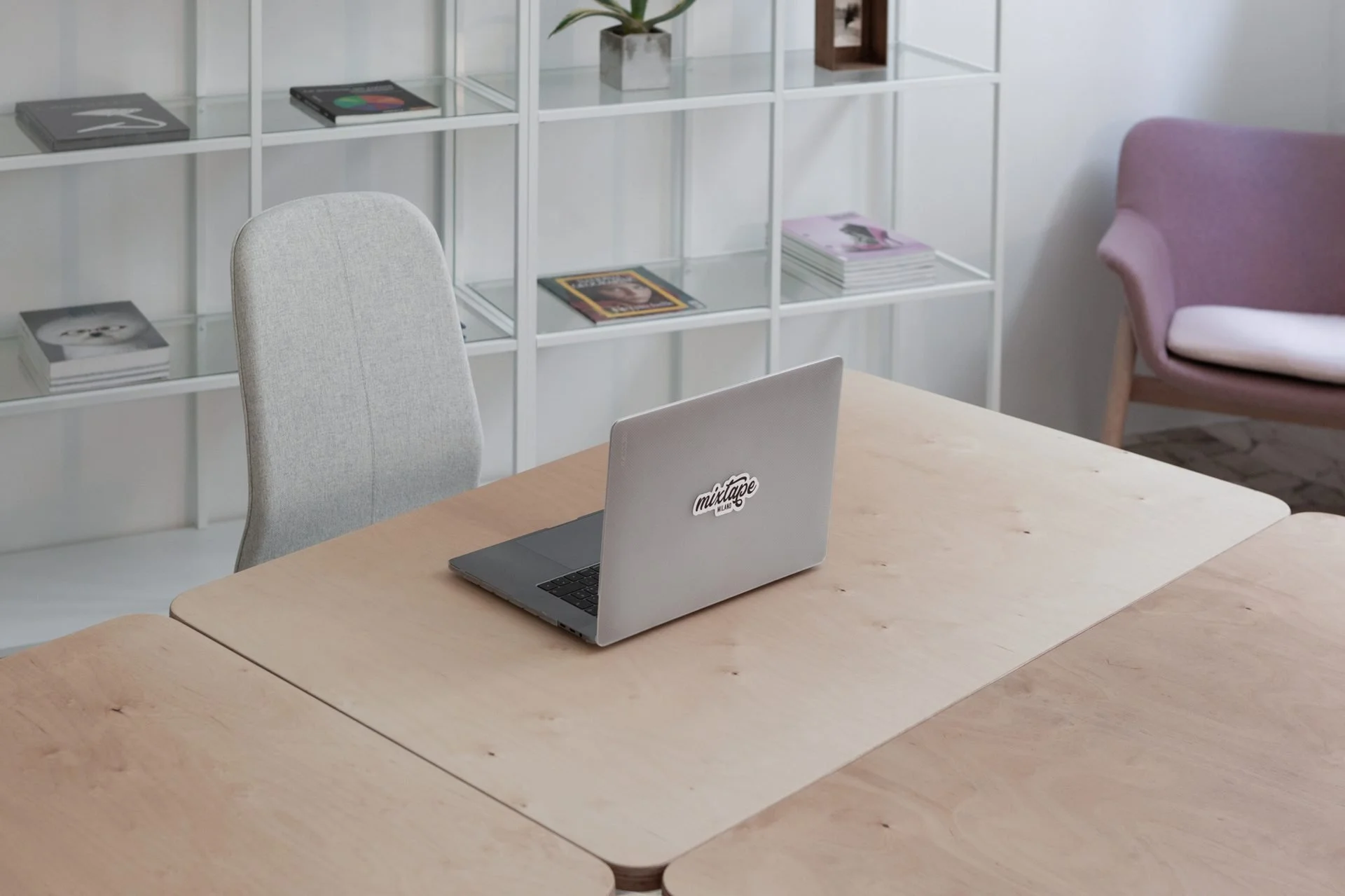 A wooden conference table with a closed silver laptop displaying a sticker that says 'meltape' in black cursive writing. In the background, there's a white shelving unit with magazines, a potted plant, and decorative boxes. A light gray upholstered chair is positioned near the desk, and a purple cushioned chair with wooden legs is partially visible to the right.