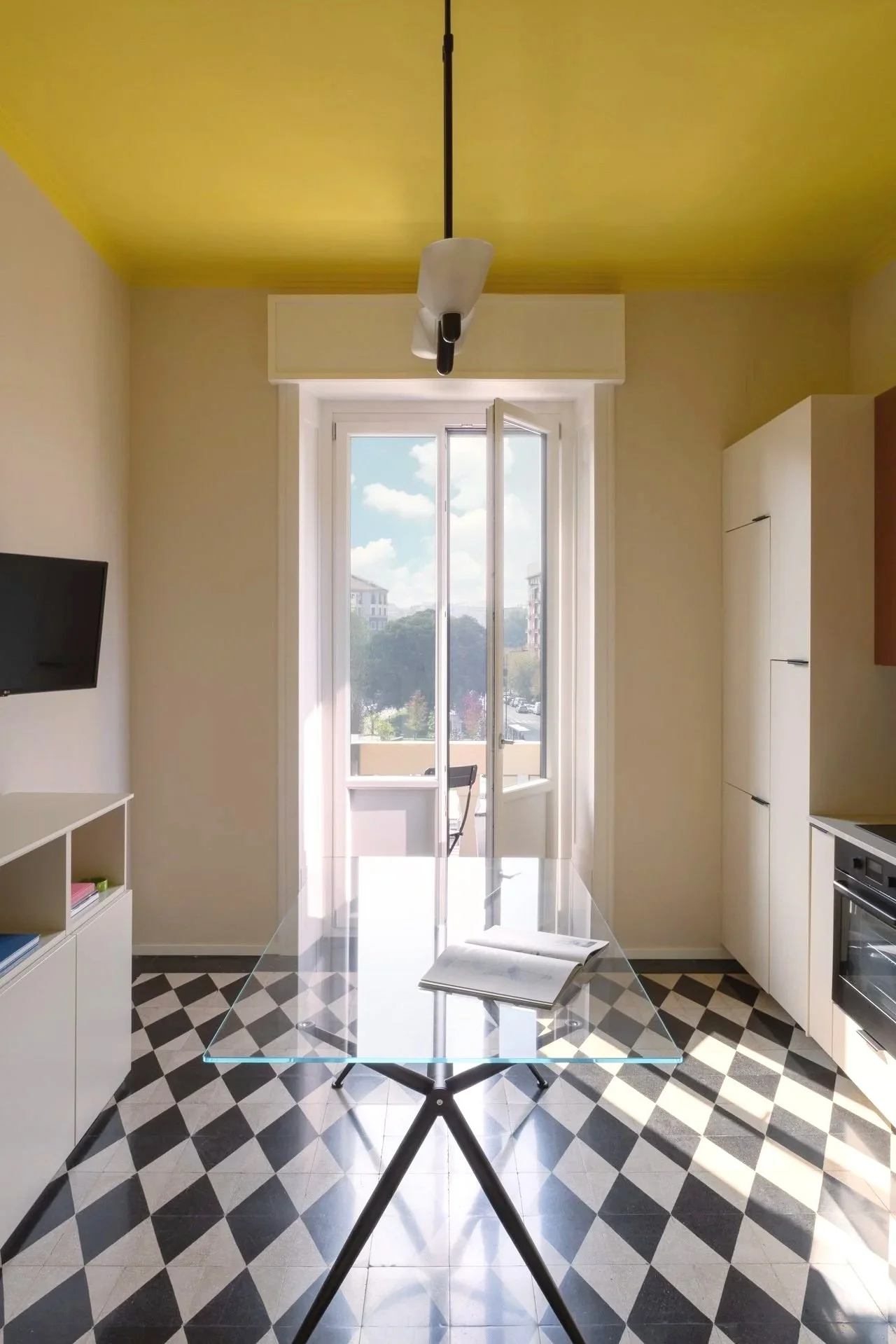 Bright room with open window showing a view of trees and buildings, white cabinets, a glass table with an open book, black and white checkered floor, and a ceiling light fixture.