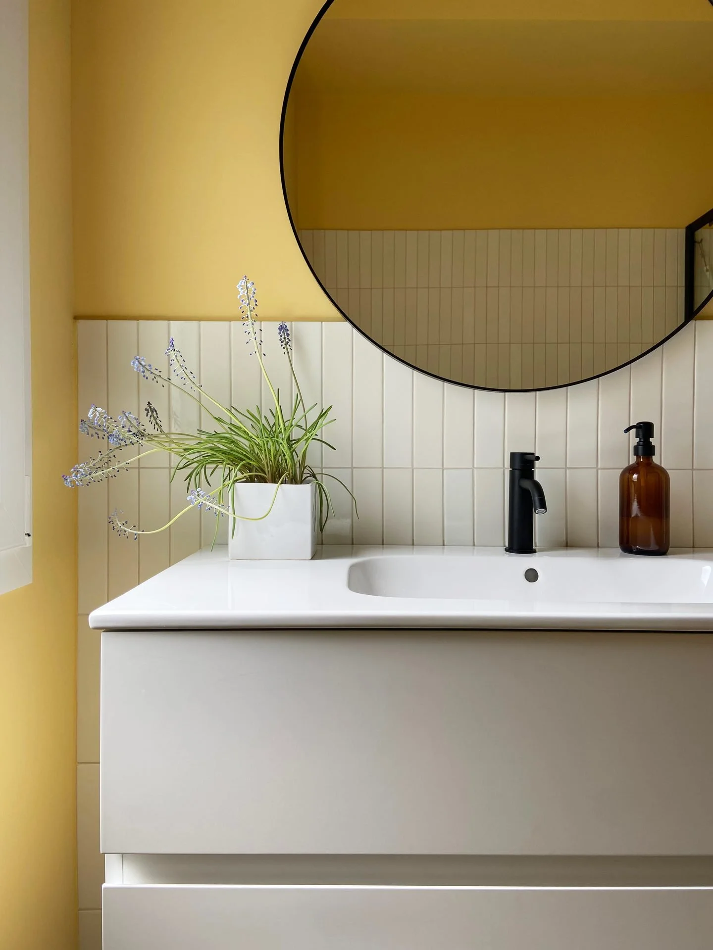 A bathroom vanity with a white countertop, a black faucet, and an amber soap dispenser. A potted plant with green leaves and purple flowers sits on the counter. A large, round mirror reflects the yellow and white tiled walls.