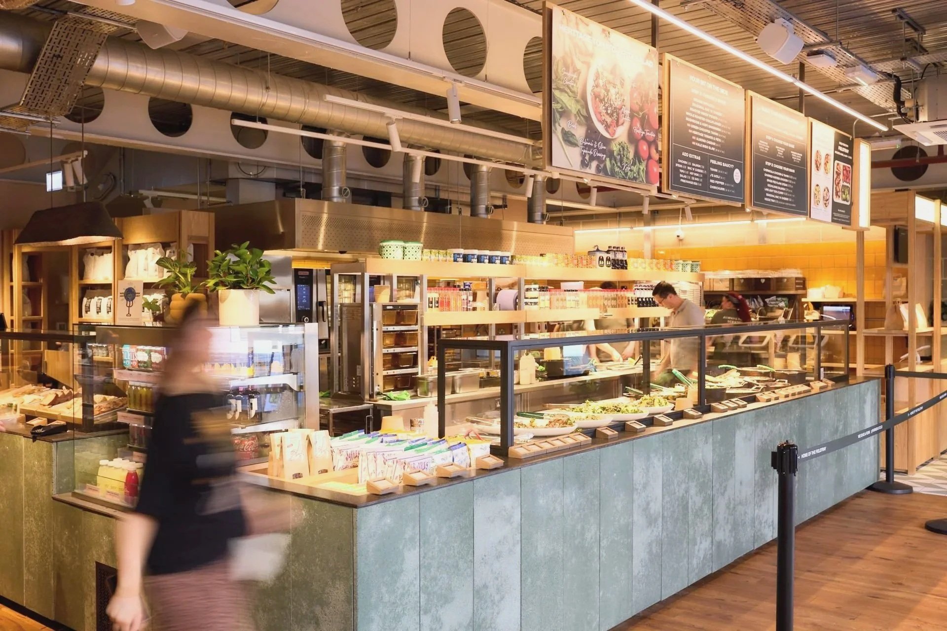Interior of a modern eatery or cafeteria with a counter, menu boards overhead, and a blurred person walking past.