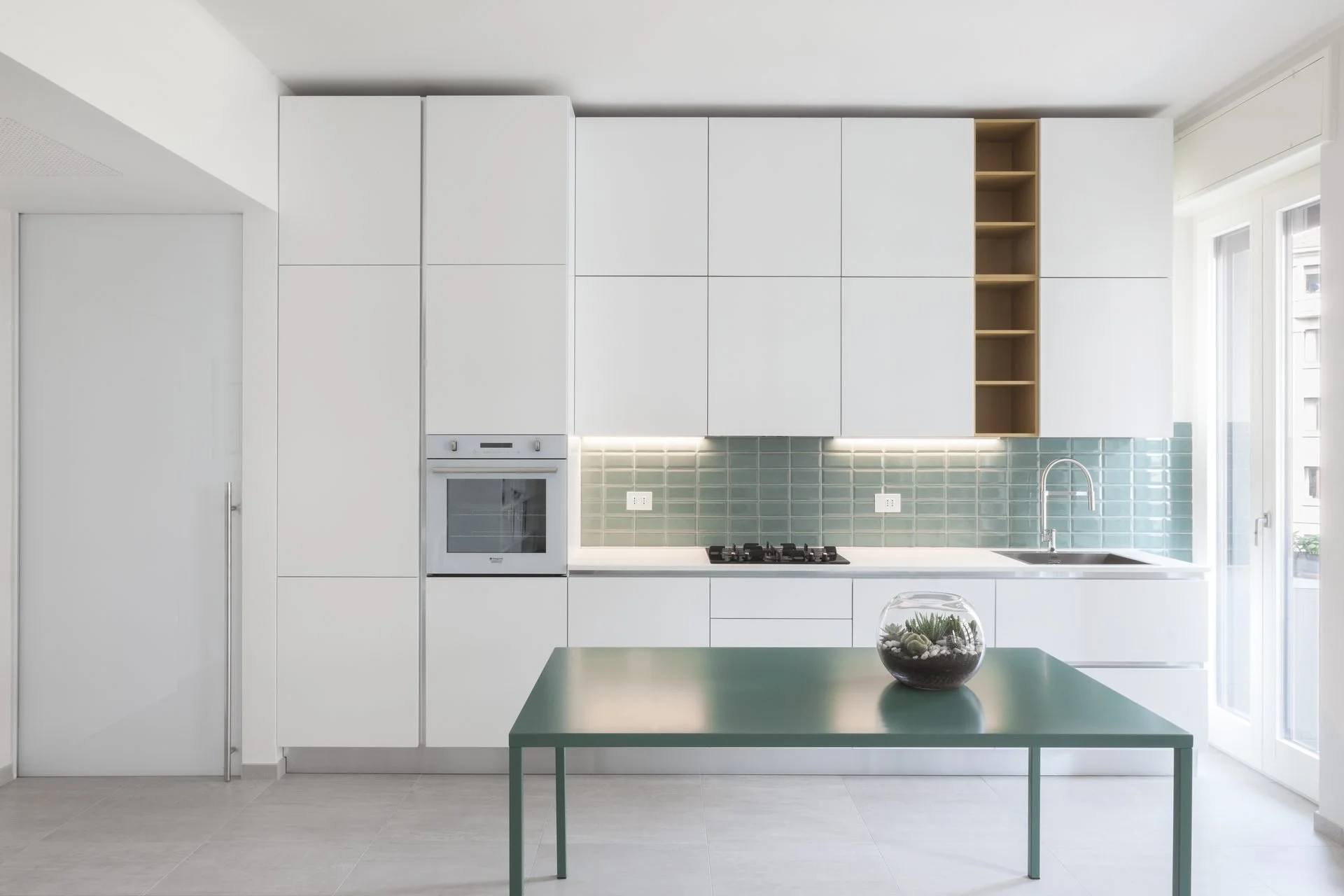Modern white kitchen with green tile backsplash, built-in oven, gas stove, stainless steel sink, and open shelving, illuminated by natural light from large windows.