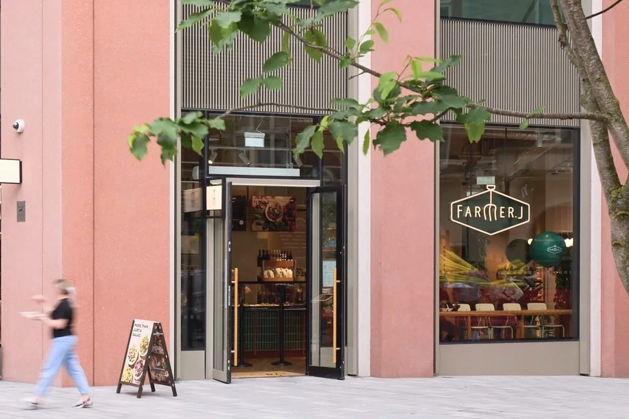 Exterior view of a restaurant called Farmer J with pink walls, glass doors, and large windows. A person is walking past, and there is an A-frame sign outside.