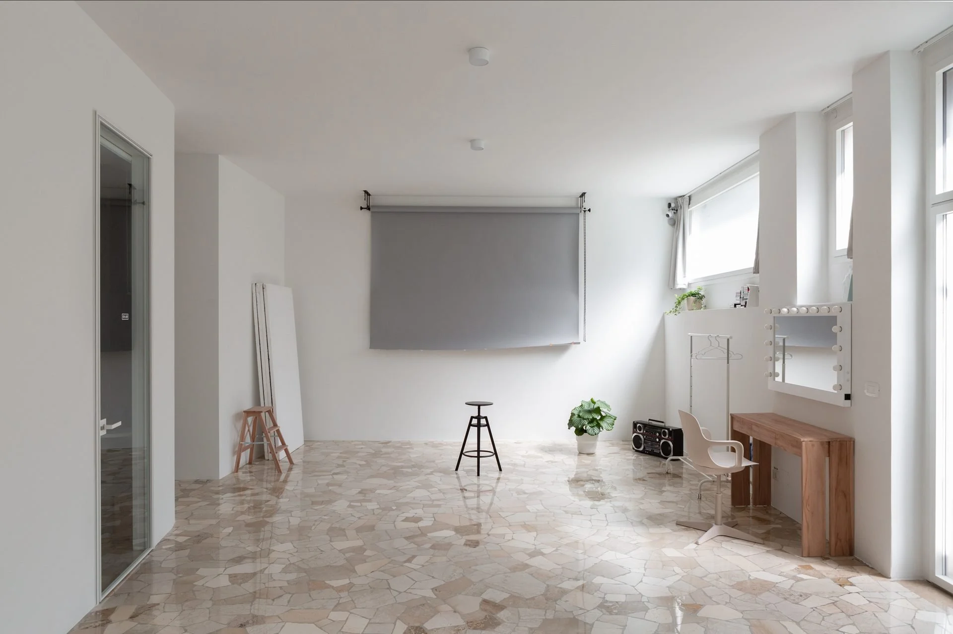 Minimalist indoor space with white walls, large windows, and tiled floor, decorated with a potted plant, a small table, chair, and a large mirror with lights.